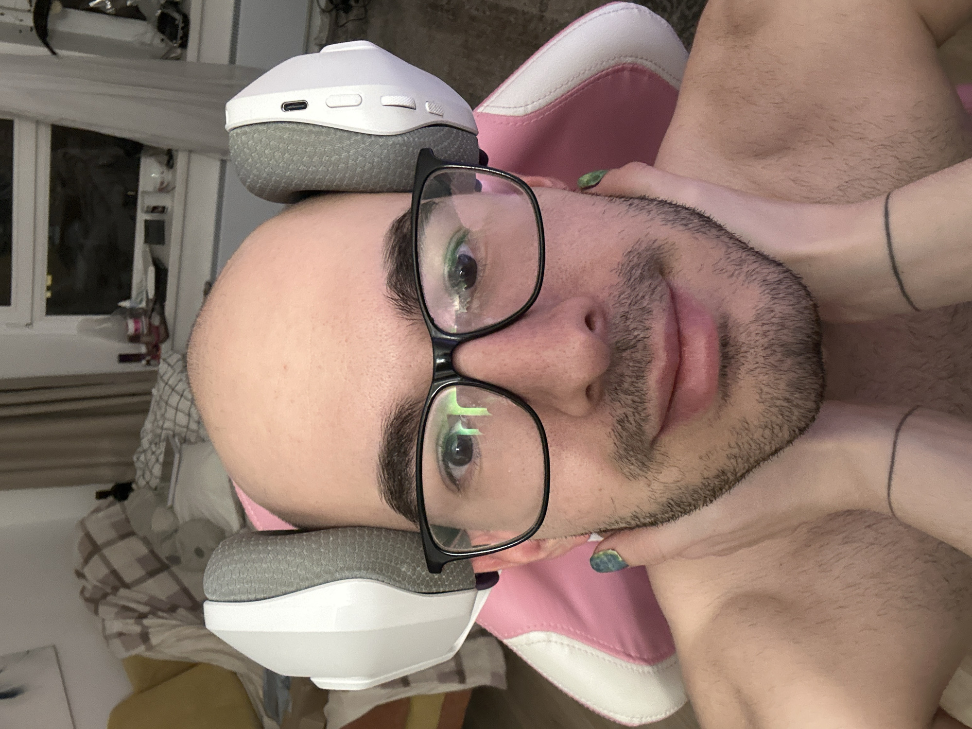 A bald, fair-skinned person wearing eyeglasses is posing for a selfie-style photograph. The person's face is centered, and they are looking directly at the camera. The person has a dark beard and mustache. The top of their head is resting on a white and gray headphone, and their neck is being held by light-skinned arms with painted fingernails. The background includes a pink and white chair and a room setting with a window. The location in the city of Moscow, Russia, cannot be determined from the image.