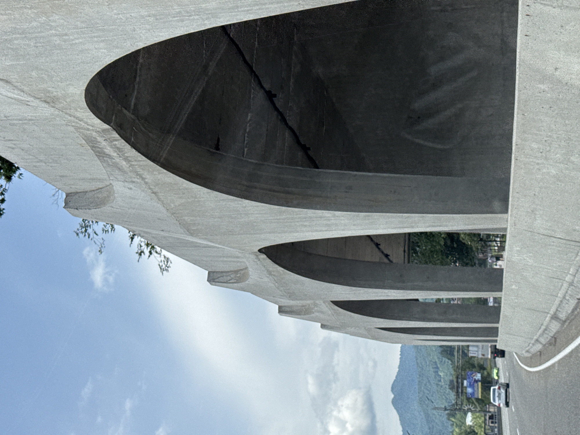 The image shows a concrete overpass with a series of arched openings. The overpass is located in Estosadok, Russia. In the foreground, a highway is visible with several cars driving on it. The overpass is made of gray concrete and has a rough texture. The arches are large and create shadows that extend into the overpass. The sky is blue with some white clouds. In the background, there are trees and mountains.