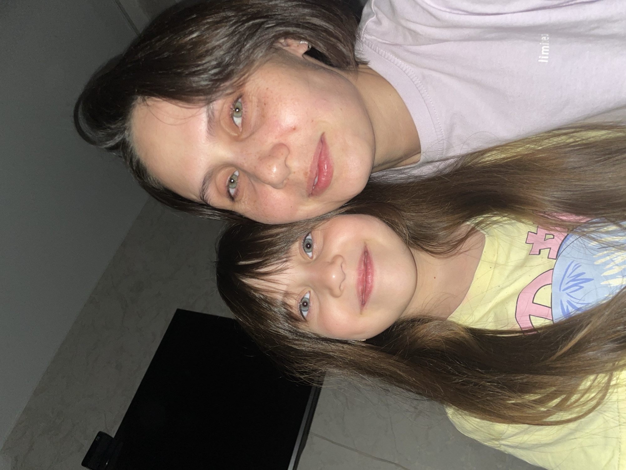 A close-up, slightly rotated photograph shows two females posing closely together. The woman has short, dark brown hair and green eyes with some freckles on her face, and is wearing a light purple t-shirt. The child has long brown hair with bangs and blue eyes, and is wearing a yellow t-shirt. The background is a dimly lit room with a dark object, possibly a television, visible in the lower left corner. No evidence indicates the location is Izhevsk, Russia.