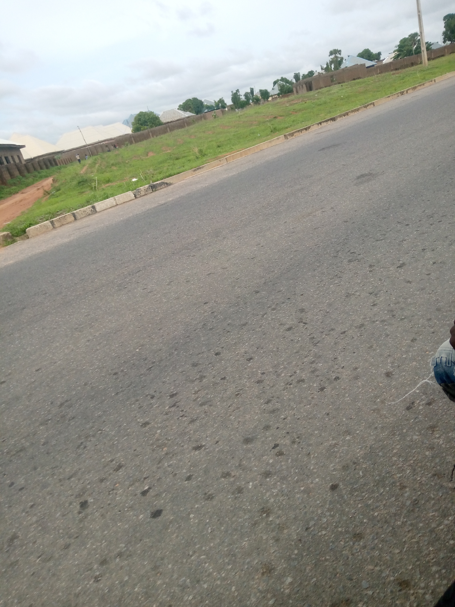 The image shows a view of a paved road in Jalingo, Nigeria, with a grassy area and some buildings on the left side of the road. The road takes up most of the frame, appearing to be a two-lane street. On the left, there is a raised grassy embankment bordered by a curb and sidewalk. Beyond the embankment, there are a few buildings under construction or recently built, some with light-colored roofing. A chain-link fence runs along the property line. A few trees are visible in the distance, and the sky is overcast. In the foreground, on the right side of the frame, a person's lower legs and feet, wearing blue jeans and a shoe, are visible. The person appears to be either standing or sitting on the edge of the road, possibly on a motorcycle or bicycle, as suggested by a thin white string extending from the person's shoe area.