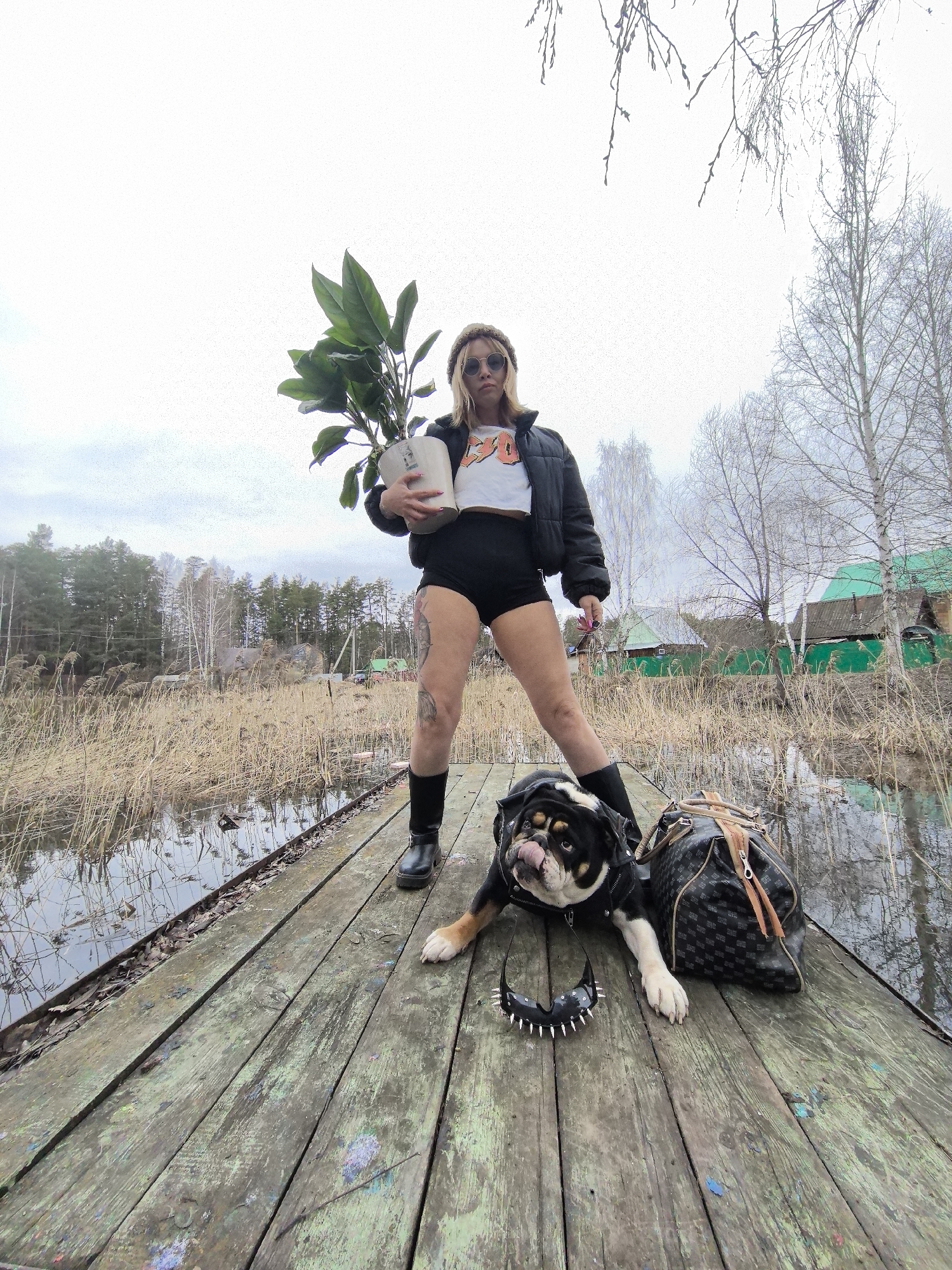 The image depicts a woman and a dog on a weathered wooden pier extending over a body of water, set in what appears to be a rural or semi-rural landscape in Noviy, Russia. The sky is overcast, and the surrounding trees are mostly bare, suggesting early spring, late autumn, or winter.
A woman stands centrally on the pier, holding a potted green plant. She is dressed in a black puffy jacket, a white crop top with an orange graphic (showing partially legible text like "EO" or "ED"), black shorts, black boots, sunglasses, and a beanie or hat. Tattoos are visible on her legs.
To her right, a bulldog-type dog lies on the pier with its tongue out, appearing relaxed. It wears a dark harness or vest. Next to the dog is a dark, patterned duffel bag with brown straps, and a black spiked collar. The wooden planks of the pier are old and discolored, with patches of green and blue paint.
The water surrounding the pier is murky, reflecting the gray sky, and is densely bordered by tall, dry reeds or grass. In the background, beyond the reeds and water, are groups of trees, including some evergreens and deciduous trees, as well as a few small, rustic houses with green roofs. The scene conveys a moment of calm amidst a natural, somewhat rugged setting.