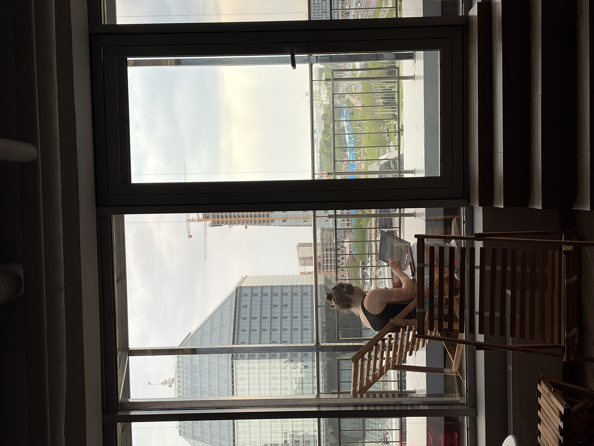 The image depicts a woman sitting on a wooden chair inside a building, working on a laptop. She is seen through a window frame, which provides a view of the city landscape of Бараўлянскі сельскі Савет, Belarus. Outside, one can see buildings, including skyscrapers, as well as greenery and infrastructure. The overall lighting suggests it is daytime. The photo is taken from an interior perspective, showing the juxtaposition of indoor activity against the urban environment outside.