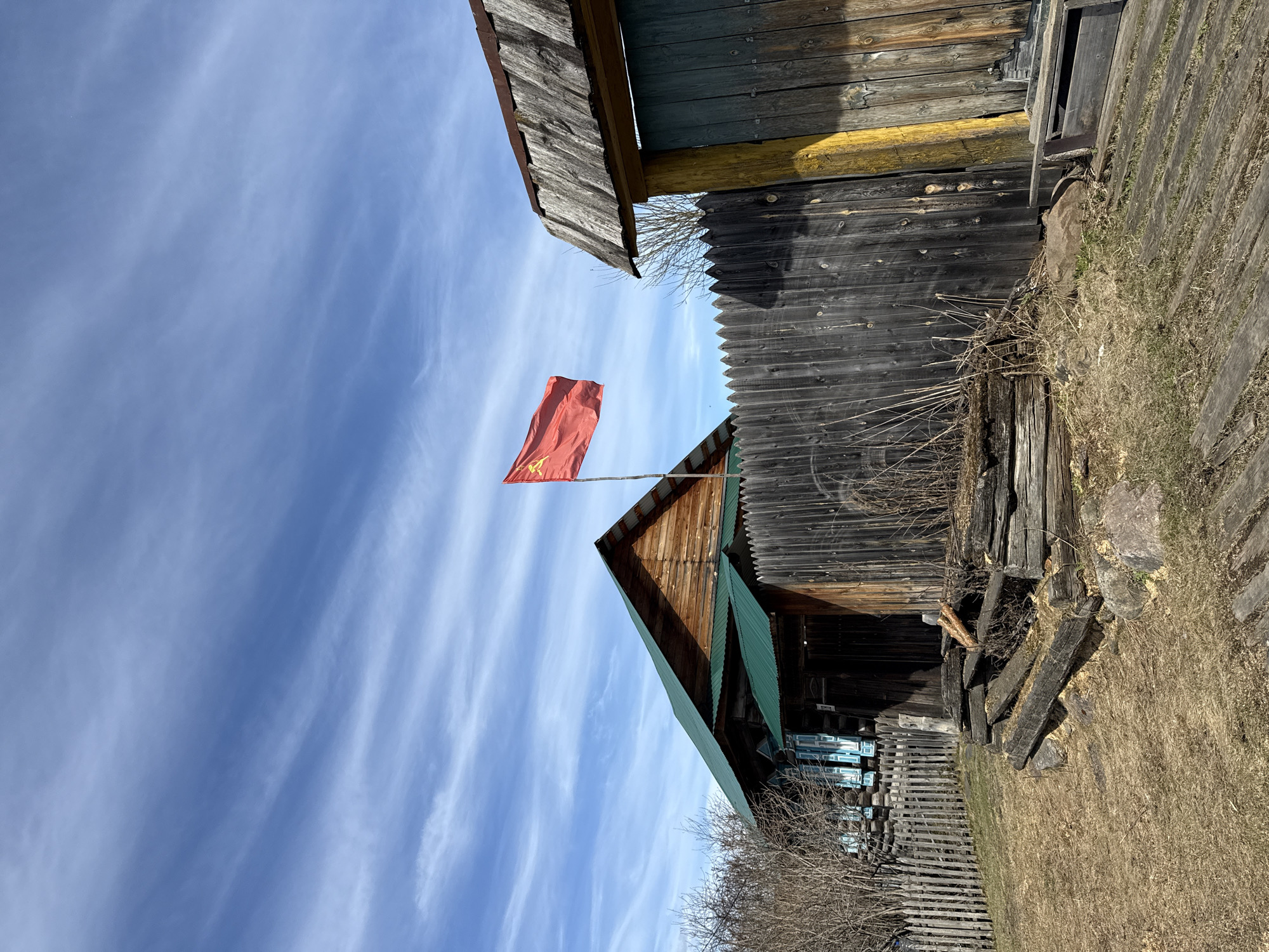 A vertical, full-shot photograph depicts a rural scene in Tyulyuk, Russia. The photograph is rotated counter-clockwise. The sky, with wispy white clouds against a blue backdrop, dominates the left side of the image.
To the right and bottom of the sky is an old, weathered wooden house and outbuildings. The house features a gabled roof, parts of which are green. A red flag with a yellow hammer and sickle emblem is attached to a pole and is perched atop the house's roof. The surrounding structures are constructed from dark, aged wood. Dry grass and bare trees and shrubs are also in the bottom and right-hand sides of the image.