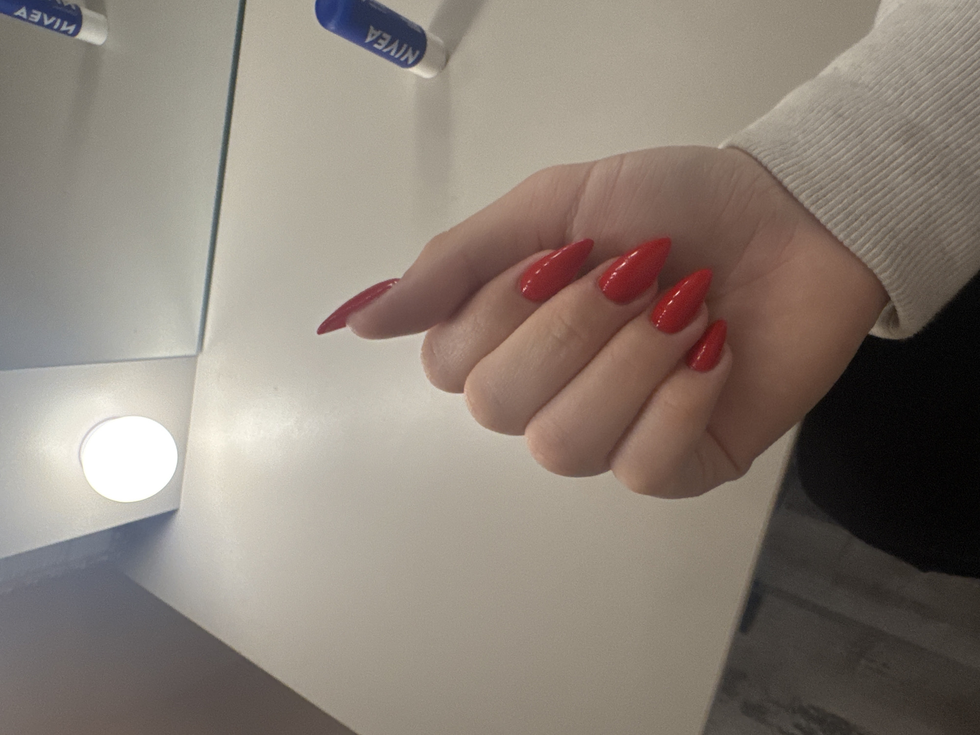 A right hand with red painted, stiletto-shaped nails is resting on a white surface. The hand is partially visible, with the wrist covered by a white knit sleeve. To the left and above the hand are two cylindrical objects, one blue and the other obscured, possibly lip balm or similar personal care products. The setting appears to be indoors, with a light source visible in the lower left corner, possibly a vanity mirror. The background is a plain white surface. The location in the city of Lehchevo, Bulgaria is not discernible.