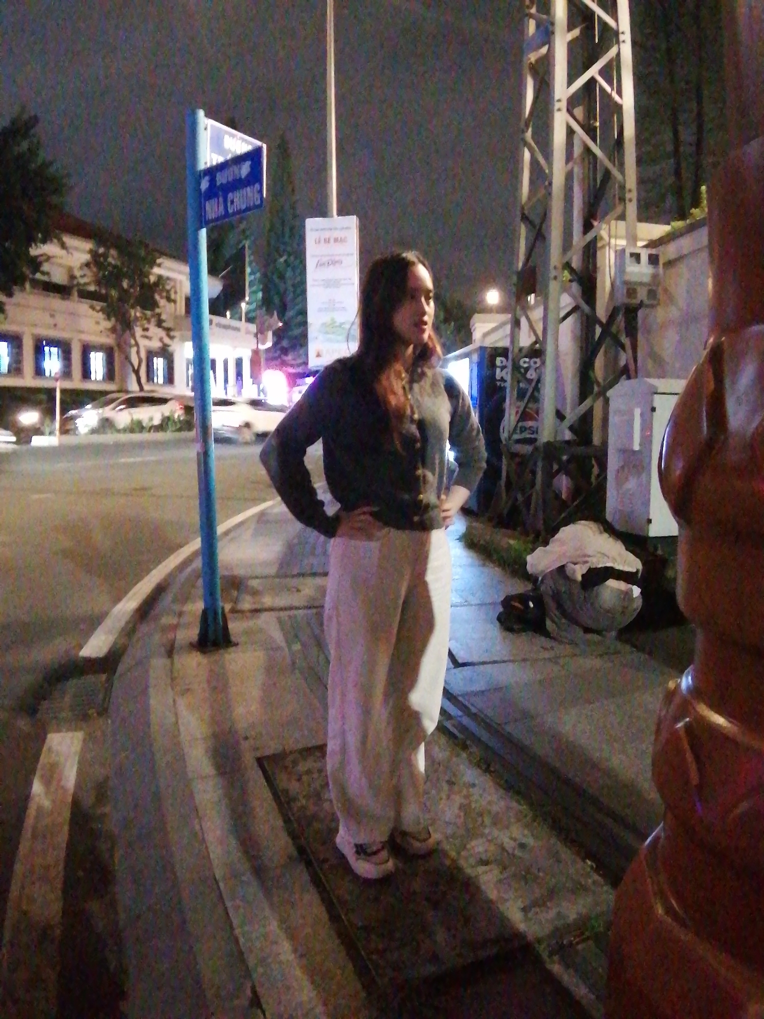 The image captures a nighttime scene on a street in Da Lat, Vietnam. A young woman with dark hair is standing on a sidewalk with her hands on her hips, facing towards the right of the frame. She is wearing a dark cardigan over a lighter top and loose-fitting light-colored pants. She is also wearing white sneakers with some visible detailing on the heel.
In the background, a blue street sign with white text is visible, indicating street names. The sign appears to read "DUONG" and "NHA CHUNG." Further down the street, a building with illuminated windows is visible. Several cars are driving on the road, some of which are blurred due to motion.
To the right of the woman, a tall metal utility pole or structure stands prominently. On the ground near the pole, there are some discarded items, including what appears to be a pile of fabric or clothing. A large, reddish-brown object, possibly a decorative element or a part of a structure, is in the foreground on the far right, partially obscuring the view. The overall lighting suggests it is nighttime, with artificial lights illuminating the street and surroundings. The environment appears urban, with a sidewalk, road, and street infrastructure.