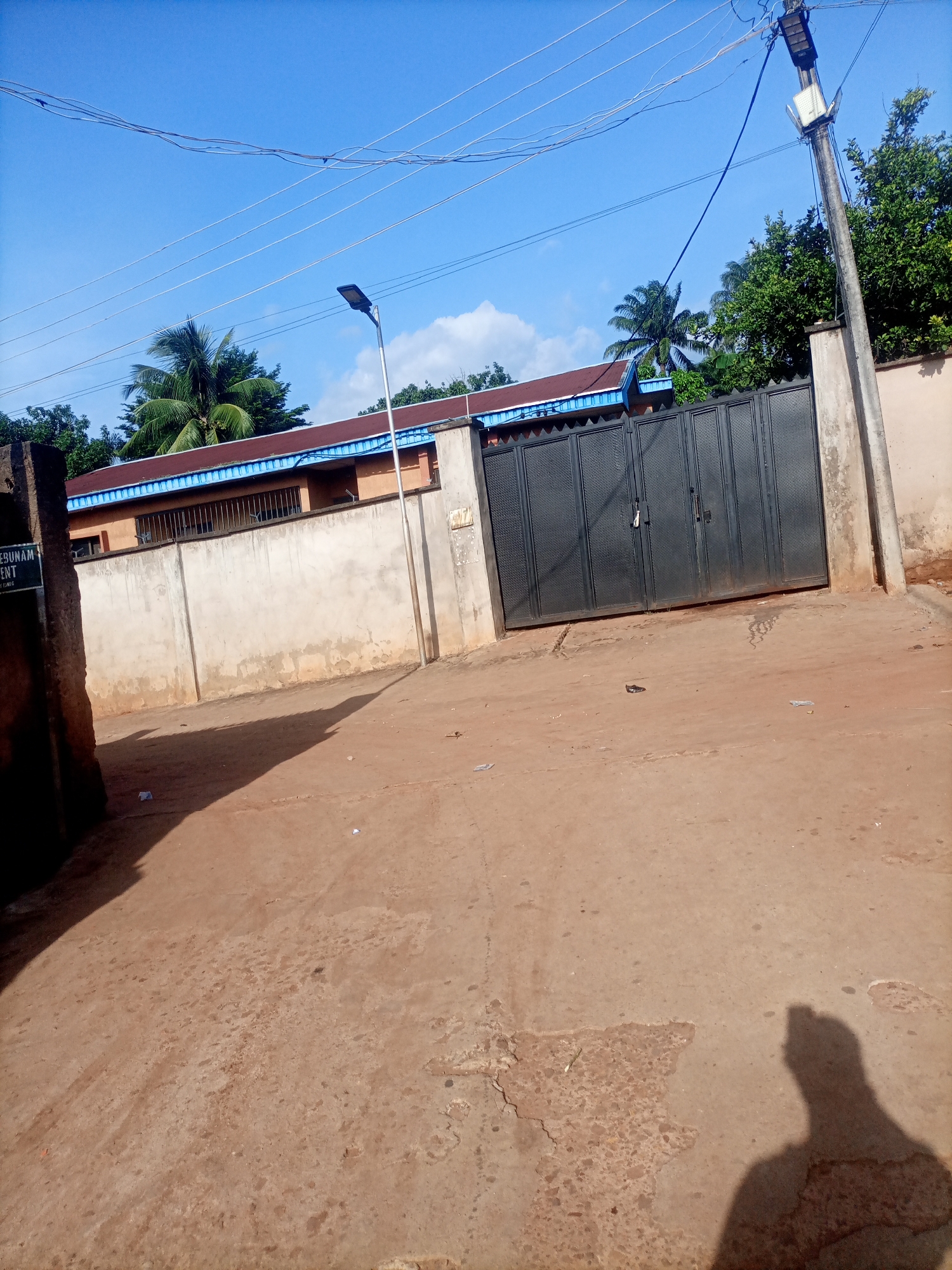 The image shows a wide shot of a street scene in Nkpor, Nigeria. The foreground is dominated by a reddish-brown, cracked concrete road, with scattered debris including small pieces of paper and a dark object that could be a piece of trash or a small animal. The shadow of a person, presumably the photographer, is cast on the right side of the road, indicating a bright, sunny day.
In the midground, a tall, light-colored concrete wall runs horizontally across the image, with a large, dark metal gate in the center. The gate is segmented into multiple panels and appears to be closed. To the left of the gate, a sign is partially visible on a concrete pillar, with some text that reads "EBUBUNAM", followed by other words that are difficult to decipher. Above the wall, the roof of a building is visible, with brown tiles and a blue trim. Palm trees and other lush green foliage are seen peeking over the roof and behind the wall, suggesting a tropical or subtropical climate.
In the background, a clear blue sky with a few scattered white clouds is visible. Several power lines stretch across the sky, supported by utility poles. A street light is mounted on one of these poles to the right of the gate, and another is visible further to the left. The overall impression is a street in a residential or commercial area on a bright, sunny day, with the presence of local flora and infrastructure characteristic of Nkpor, Nigeria. No specific activity or event is clearly depicted, but the scene suggests an ordinary day.