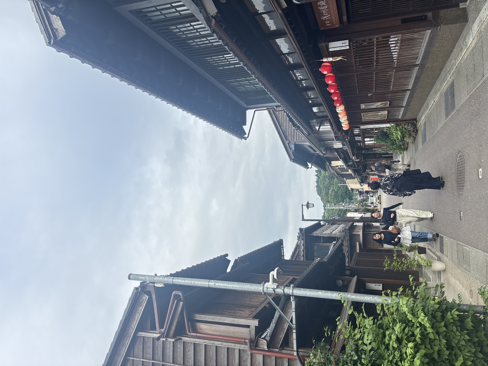 An overhead view captures a traditional street scene in Kanazawa, Japan, under an overcast sky. The image is oriented vertically, showing the sky at the top and bottom with buildings on either side, as if looking up a narrow street from a low angle. When viewed correctly, the perspective is down a historic street flanked by dark-stained wooden buildings with traditional tiled roofs and intricate latticework.
On the right side of the street, a row of red and white lanterns with floral patterns hangs from the eaves of a building, beneath a sign with Japanese characters and partially visible English text that might read "SAMURAI-GASHIRUYA-EN". Below the lanterns, two framed pictures are displayed on the wooden facade. The left side of the street features similar traditional wooden architecture, with prominent drainage pipes and gutters. A utility pole with a mounted camera is visible in the foreground on this side.
Several people are walking along the paved street, moving away from the viewer. One individual in a dark, possibly traditional, garment walks in the center. Behind them, two women in casual attire – one in a black top and white pants, the other in a black top, blue jeans, and a white ruffled undershirt, carrying a light brown bag – are visible. Green foliage, including bushes and potted plants, lines the sidewalk. In the distance, more buildings and a tree-covered hillside are visible under the muted light of the cloudy day. The overall atmosphere suggests a quiet, historic district.