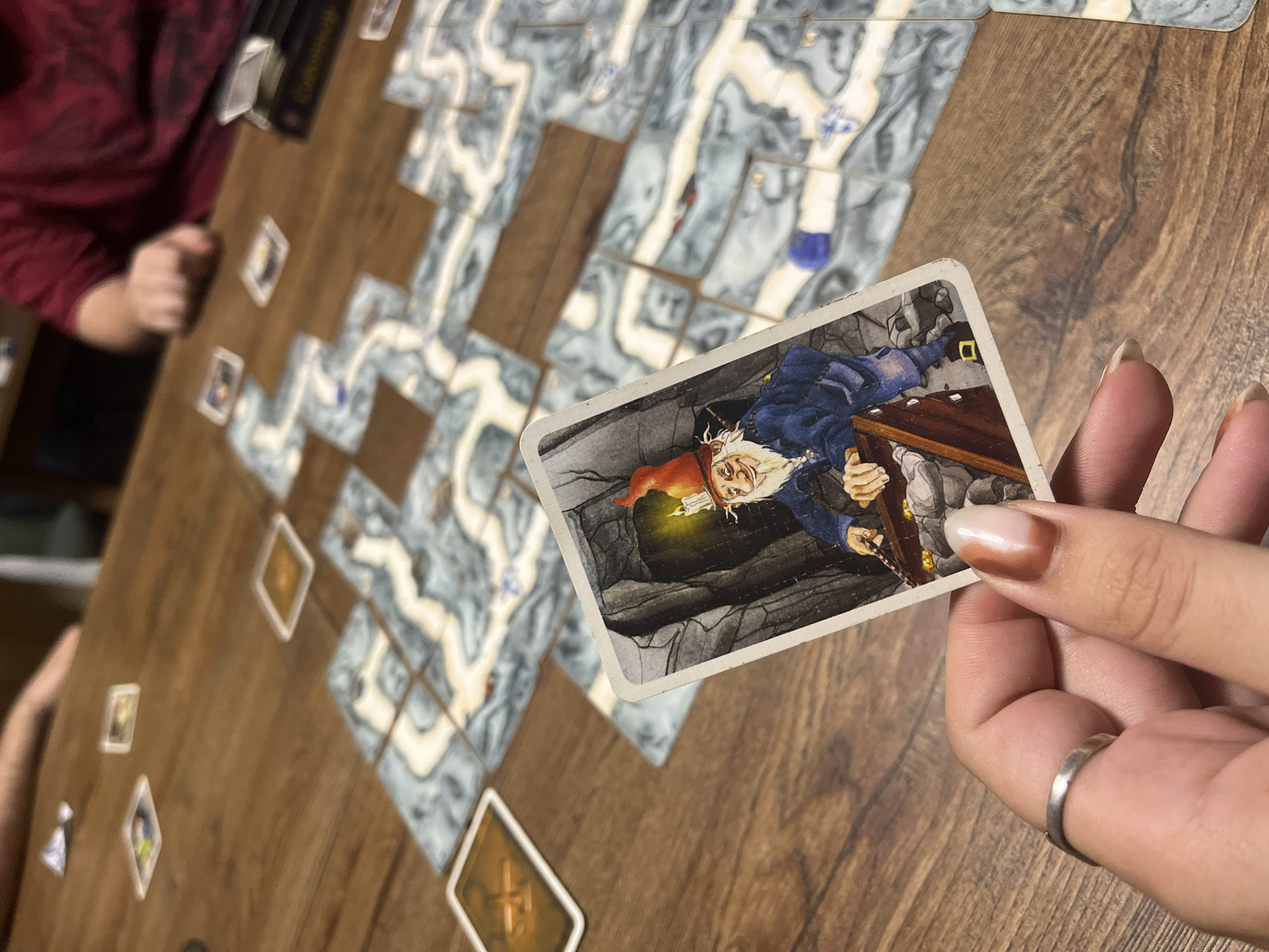 A close-up image shows a person holding a game card. The card depicts a miner with a red cap and a lamp digging in a mine. On a wooden table are several tiles depicting a meandering cave system. Other game cards are scattered on the table. The image lacks discernible characteristics to associate it with Varna, Bulgaria.