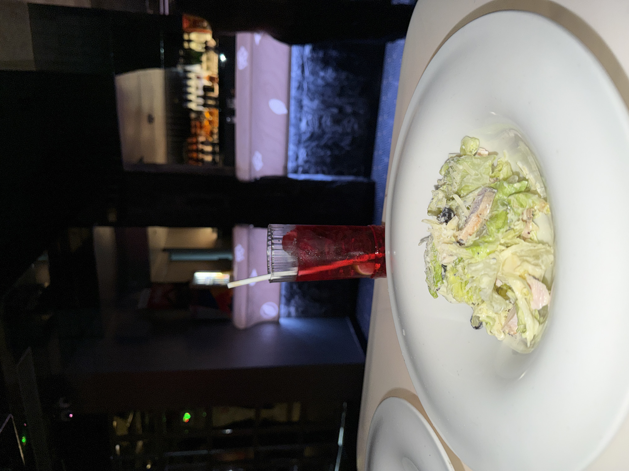A high-angle, low-light photograph captures a dining setting within a modern establishment identified as New Delhi, India. In the immediate foreground, a white, wide-rimmed bowl contains a salad composed of chopped green lettuce, pieces of light pink meat (likely poultry or seafood), and a creamy dressing, topped with grated white matter. To the left, a portion of another white plate is visible. Behind the salad, a tall, clear, ribbed glass holds a red liquid, ice cubes, and what appear to be red fruit garnishes, with a white straw inserted. The table surface is light beige. The mid-ground and background are out of focus, revealing a dark environment. A bar counter with white panels featuring circular cutouts extends across the mid-background, illuminated by blue light strips at its base. Above the counter, shelves are stocked with numerous bottles, some reflecting light. On the far right, a dark, indistinct silhouette of a person is present. On the far left, blurred green and pink lights are visible. The overall scene depicts a prepared meal and drink in an ambient, dimly lit restaurant or bar setting.