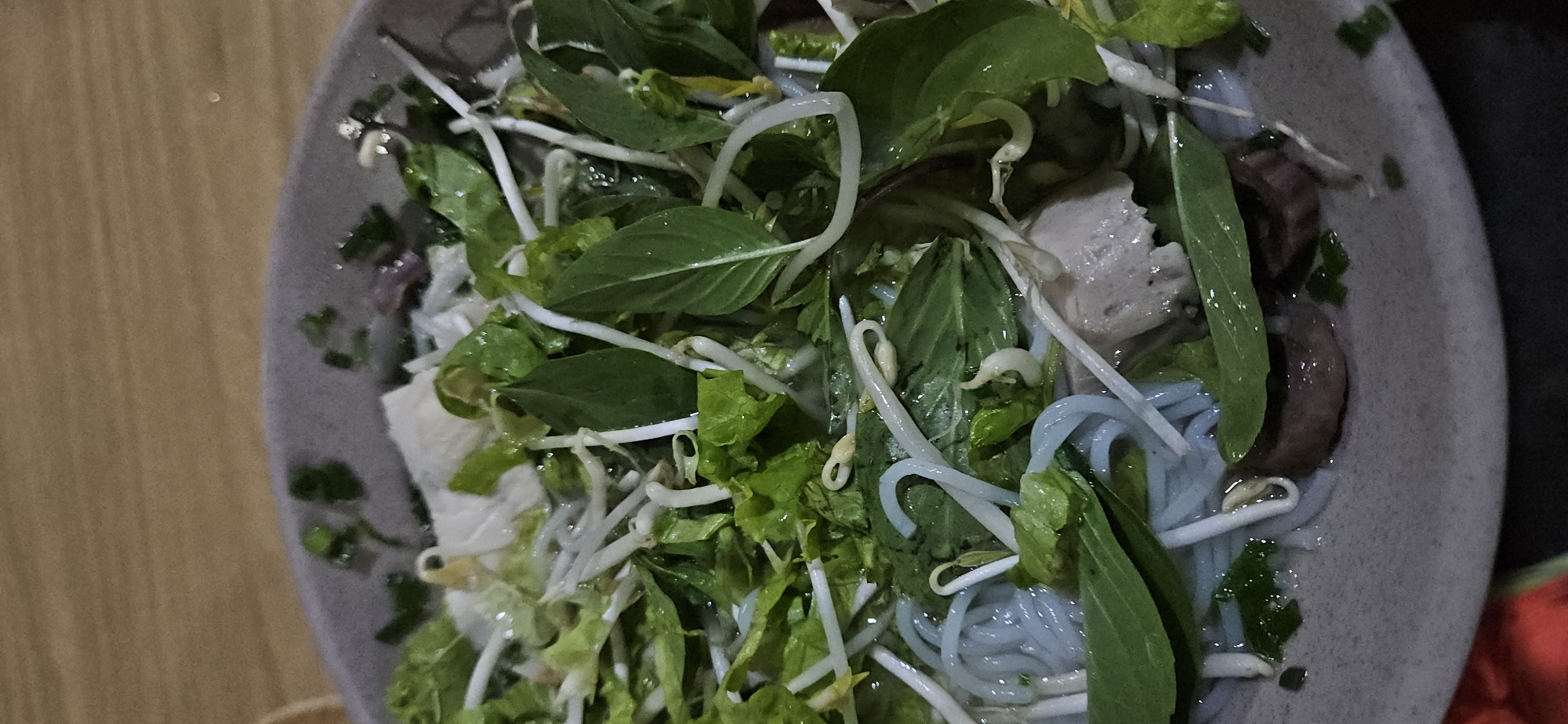 An overhead close-up shot captures a bowl of Vietnamese noodle soup or salad, likely a regional specialty from Cam Đức, Vietnam. The bowl, which appears grey or light brown, is filled with a vibrant mix of ingredients. Prominently featured are light blue or greyish thin noodles, suggesting a unique variety like Bún Đậu Mắm Tôm or a similar rice vermicelli dish.
A generous topping of fresh green herbs, including what looks like basil leaves and lettuce, along with numerous white bean sprouts, covers the surface of the dish. Beneath these greens, various protein components are visible: light-colored, somewhat irregular chunks that could be tofu or a type of processed meat, and darker, brownish pieces which might be mushrooms or a different meat. Chopped green onions or chives are also scattered throughout.
The bowl rests on a light brown wooden surface, possibly a table, which is visible on the left side of the frame. The lighting is soft and even, suggesting an indoor setting. No specific time of day or weather can be inferred. No people or text are visible in the image. The composition focuses tightly on the bowl, highlighting the textures and freshness of the ingredients.
