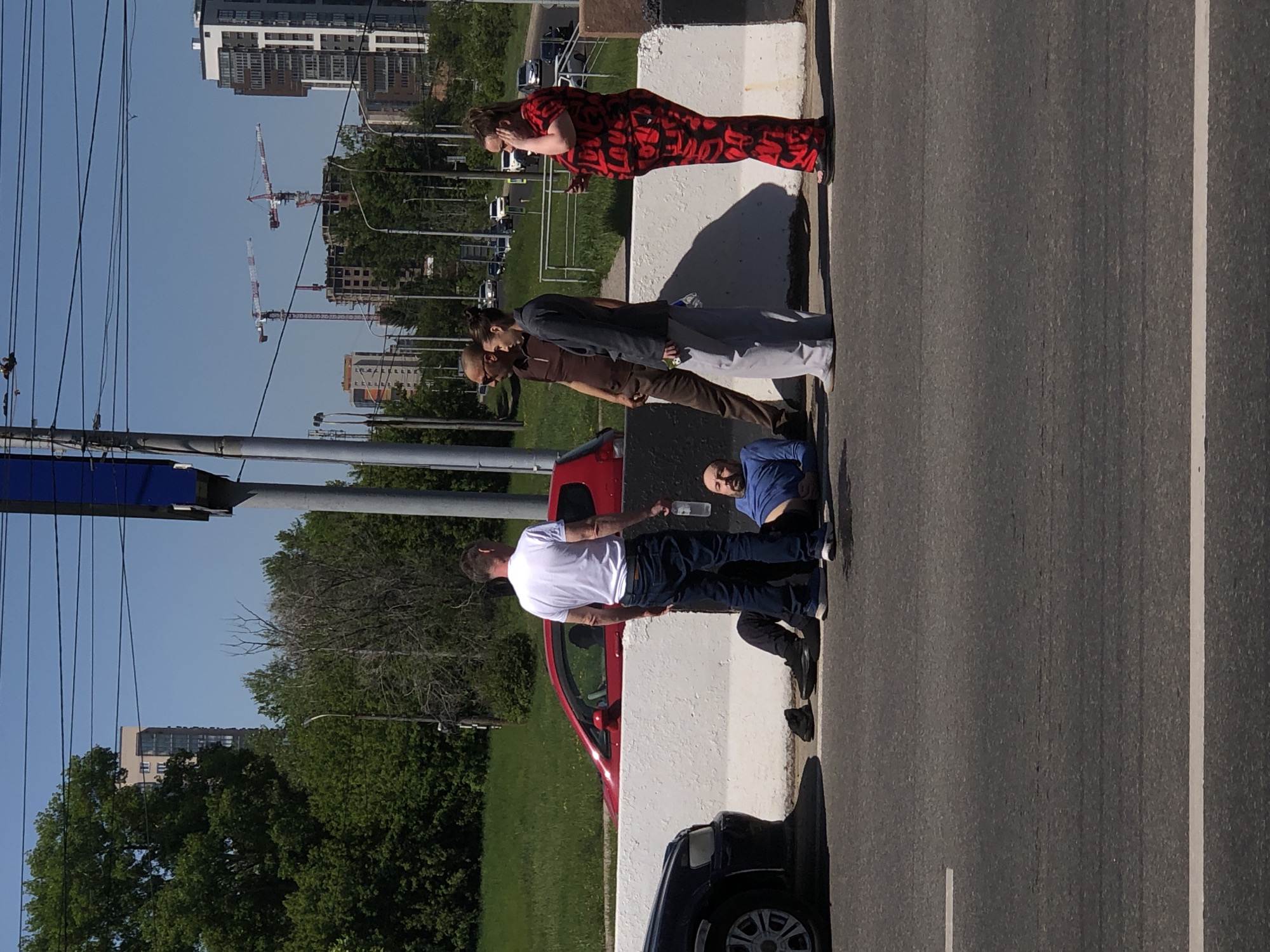 The image captures an urban street scene in Krasnoyarsk, Russia, under clear and sunny daytime conditions. Several individuals are gathered along a concrete barrier separating a pedestrian area from a multi-lane asphalt road.
In the immediate foreground, a man in a white t-shirt and dark jeans stands beside a red hatchback car, holding a transparent bottle. Adjacent to him, another man in a blue shirt is crouched or sitting on the ground. Further along the barrier, a man in a brown shirt stands with another man in a grey jacket and light pants, who appears to be holding a small can or bottle. A woman in a distinctive red and black patterned outfit stands nearby, seemingly adjusting her hair or ear. Additional people are partially visible further down the road, and a black SUV is parked near the barrier.
The background reveals a bustling city environment. Numerous tall buildings, ranging from residential to commercial structures, rise against the clear blue sky. At least two large construction cranes are prominent in the distance, indicating ongoing urban development. Lush green trees and grassy areas are visible beyond the road and barrier. The people appear calm, standing casually, suggesting they are either waiting or observing their surroundings. No discernible text is visible on signs, clothing, or objects.