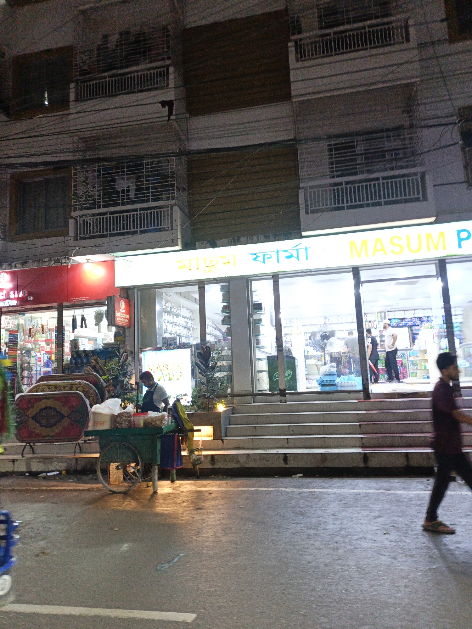 The image depicts a street scene at night in Dhaka, Bangladesh, in front of a building with multiple floors. A brightly lit storefront, identified as "MASUM PHARMA," is prominently displayed on the ground floor, featuring large glass windows showcasing products. To the left of the pharmacy, another storefront with red signage is partially visible.
In front of the buildings, a street vendor is operating a cart laden with goods, possibly food items. The cart has a green body and two wheels, with a man in a dark shirt attending to it. A few people are seen walking on the street and entering the pharmacy. The street is wet, suggesting recent rain, and the lighting indicates it is nighttime. The building itself has balconies with railings, and various wires crisscross above the street. The overall atmosphere is that of a bustling urban environment at night, with commercial activity in full swing.