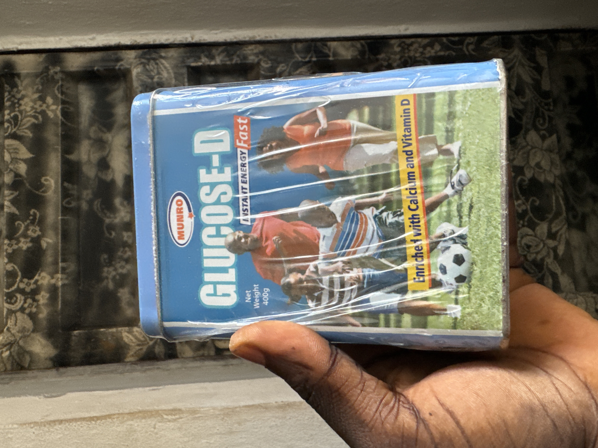 A person's thumb and index finger are holding a blue tin of "Glucose-D" drink powder. The tin is wrapped in clear plastic. The tin has an image of a family playing soccer on a grassy field. The text on the tin indicates "Instant Energy Fast" and "Enriched with Calcium and Vitamin D". The background of the image is a wall with a patterned wallpaper. The scene is indoors. It is not possible to determine the specific location within Awka, Nigeria from the image.