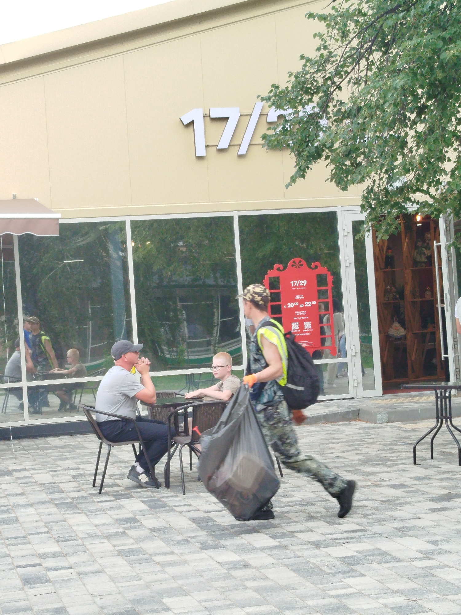 A man in camouflage pants and a yellow vest walks past a cafe in Zavodoukovsk, Russia, carrying a large black trash bag. He is walking on a paved outdoor seating area in front of a building with large windows displaying merchandise on shelves. Two men and a boy are seated at a table inside the cafe, reflected in the window. Another man is standing behind them. The building has "17/29" displayed in large white letters on its facade. A red sign with text in Cyrillic is visible in the window. The ground is paved with grey and beige square tiles.