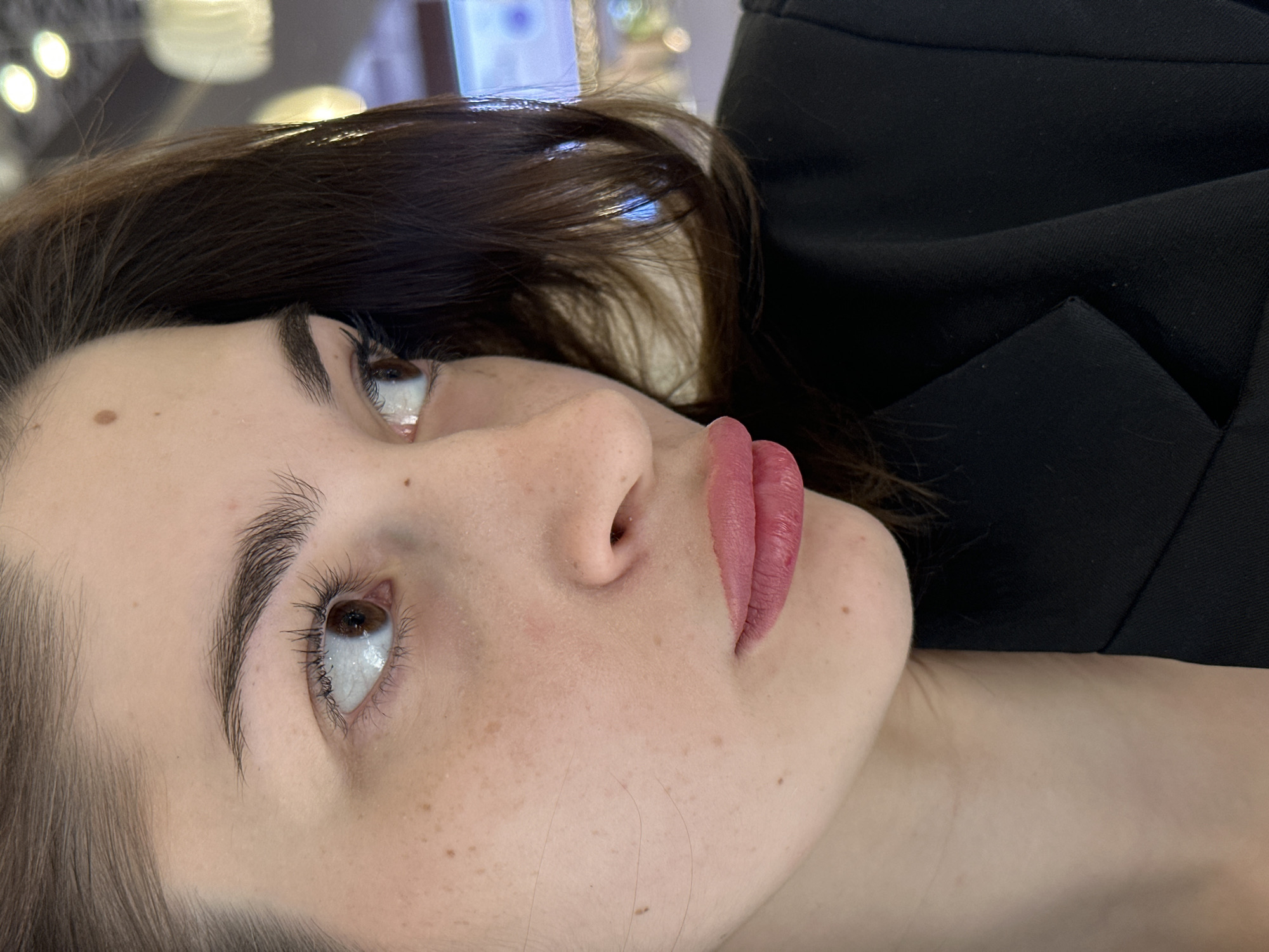 A close-up portrait shows a young adult female from the chest up, angled to her left. She has fair skin with scattered freckles across her nose and cheeks. Her right eye, with a brown iris and wide-exposed sclera, looks upward and to the right. A dark mole is present on her forehead above her right eyebrow. Her full lips are puckered forward and coated with pink lipstick. Dark brown hair frames the right side of her face. She is wearing a dark, likely black, tailored jacket or blazer. The background is blurred, showing various out-of-focus light sources including warm circular bokeh, a distinct bell-shaped light fixture on the upper right, and a horizontal blue light source, consistent with an indoor public establishment such as a shopping mall or cafe. No specific landmarks or signage are present to identify a precise location within Yekaterinburg, Russia.