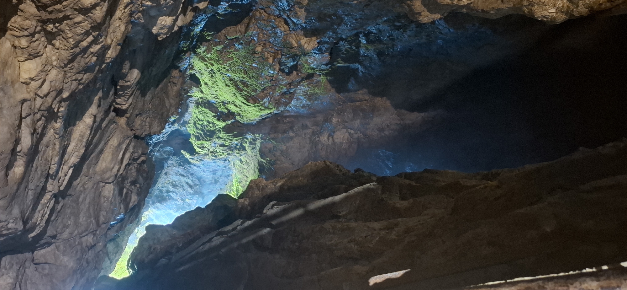 The image depicts the interior of a cave, likely within the Trigrad Gorge area of Bulgaria. The perspective is looking upwards and into the depths of the cave. A prominent feature is the jagged, rocky ceiling of the cave, with layers of stone illuminated by an unseen light source from above. Patches of vibrant green moss cling to some of the rock formations on the right side of the image, contrasting with the earthy tones of the stone. Light beams penetrate from an opening in the cave ceiling, illuminating mist or dust particles in the air, creating an ethereal glow. The cave floor is not visible, but the darkness suggests a deep chasm or passage extending downwards. The overall impression is one of natural beauty and a sense of immense scale within the cave system.