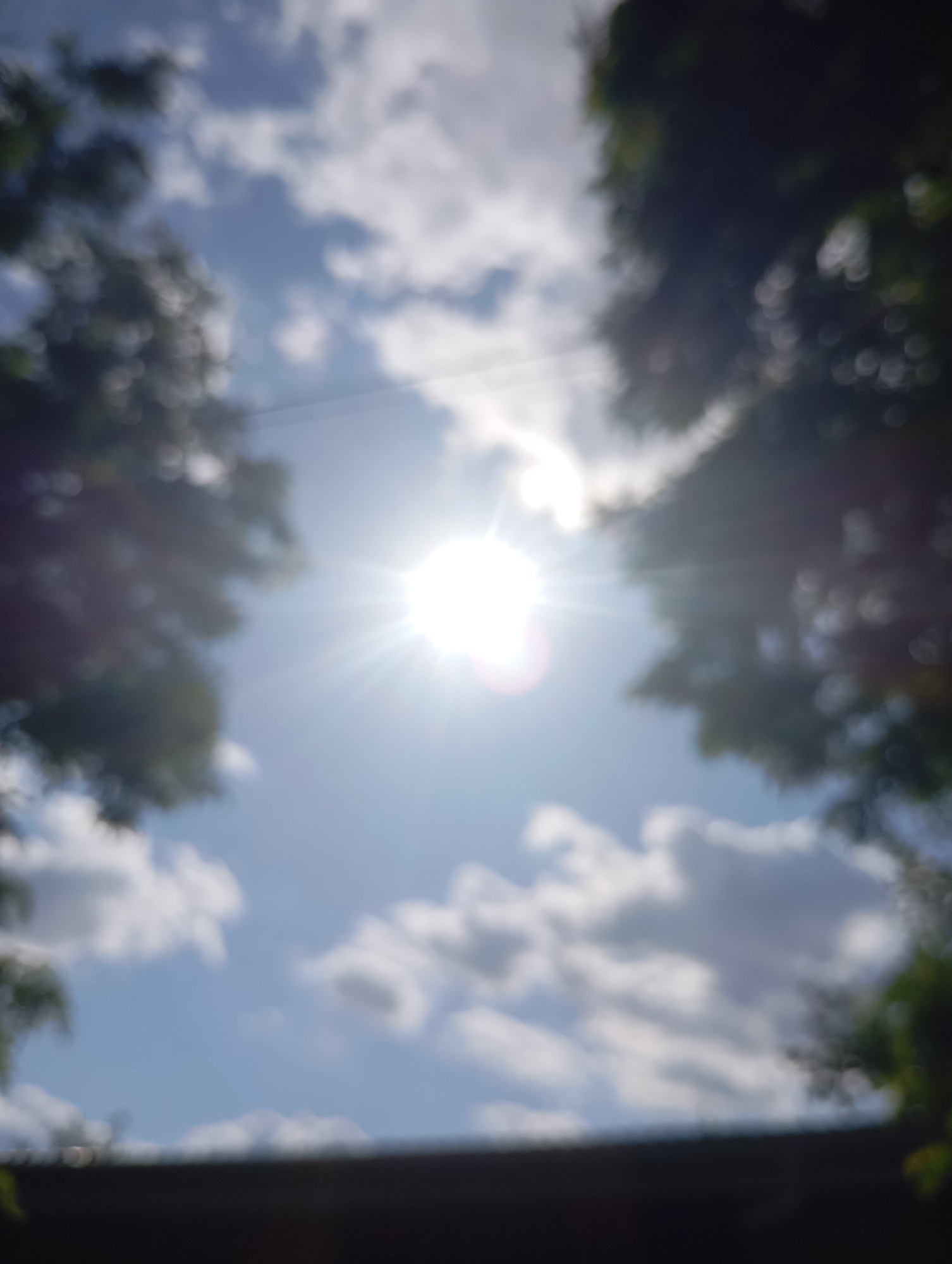 The image, taken in Sozonivka, Ukraine, is a blurry, overexposed photograph of the sky on a sunny day. The sun is the brightest element, positioned centrally and radiating light in a starburst pattern. Fluffy white clouds are scattered across a bright blue sky. Dark, out-of-focus foliage frames the top edges of the image, suggesting trees on either side. The bottom of the frame is dominated by a dark, blurry horizontal band, possibly representing a roofline or a dark surface. No people or specific structures are discernible due to the lack of focus and overexposure.