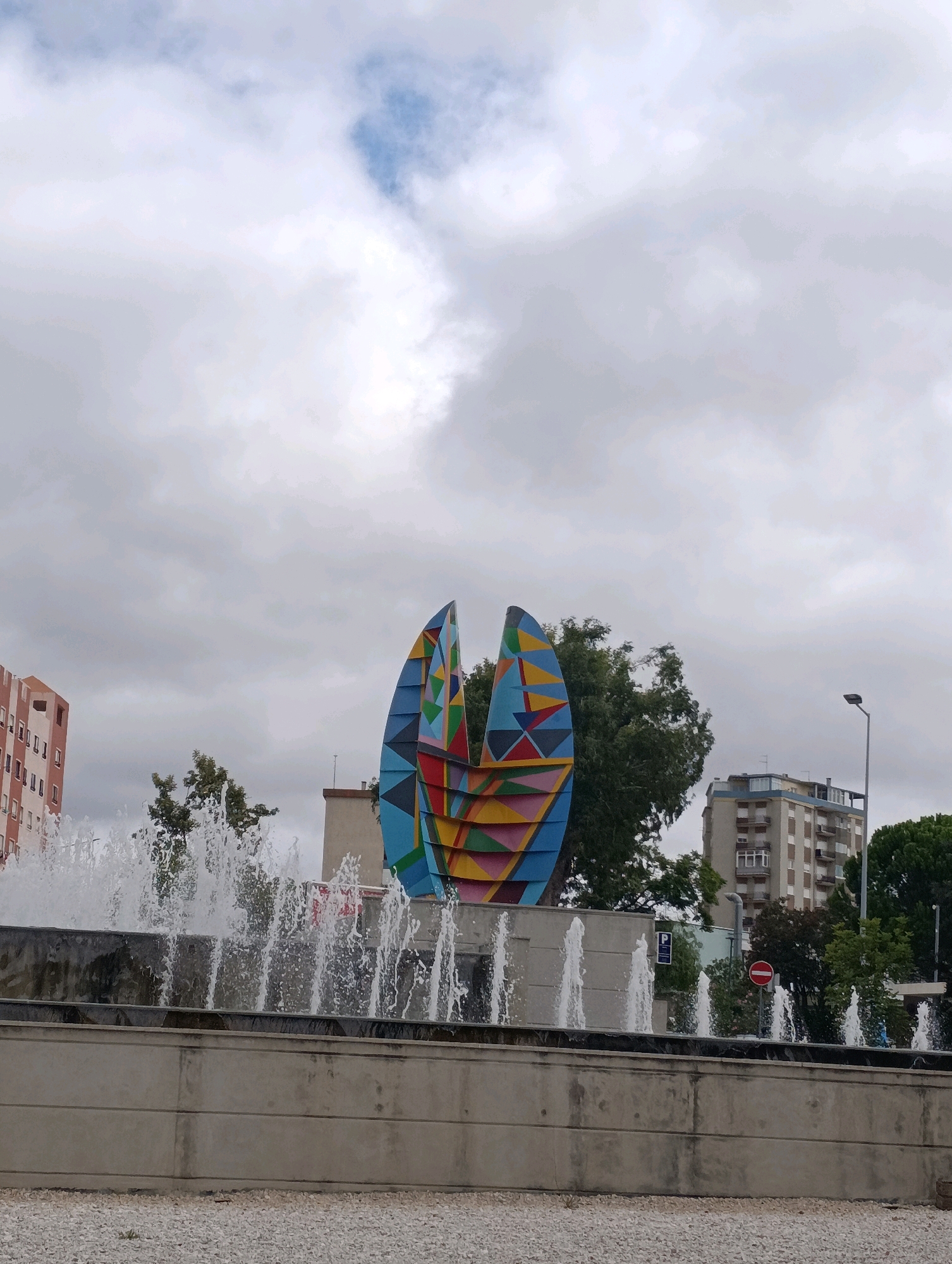 The image captures an outdoor scene in Setúbal, Portugal, likely in an urban public space. In the foreground, a multi-tiered fountain with numerous jets of water is visible. The fountain is constructed with concrete walls. Behind the fountain, a large, abstract sculpture dominates the center of the frame. This sculpture is composed of two large, curved panels painted with a vibrant, geometric pattern featuring triangles and diamonds in various colors, including blue, yellow, red, and pink.
To the left of the sculpture, a tall, multi-story building with an orange-tinted facade and visible windows stands. To the right of the sculpture, a cluster of lush green trees partially obscures another building. Further to the right, a taller, light-colored residential building with multiple balconies is visible, along with a street lamp. In the background, a cloudy sky with patches of blue completes the scene. The ground in front of the fountain is covered with small white gravel. Traffic signs, including a parking sign and a red circular no-entry sign, are present in the middle ground. A few indistinct figures are visible near the fountain on the right side of the image. The overall impression is that of a public square or park area within the city.