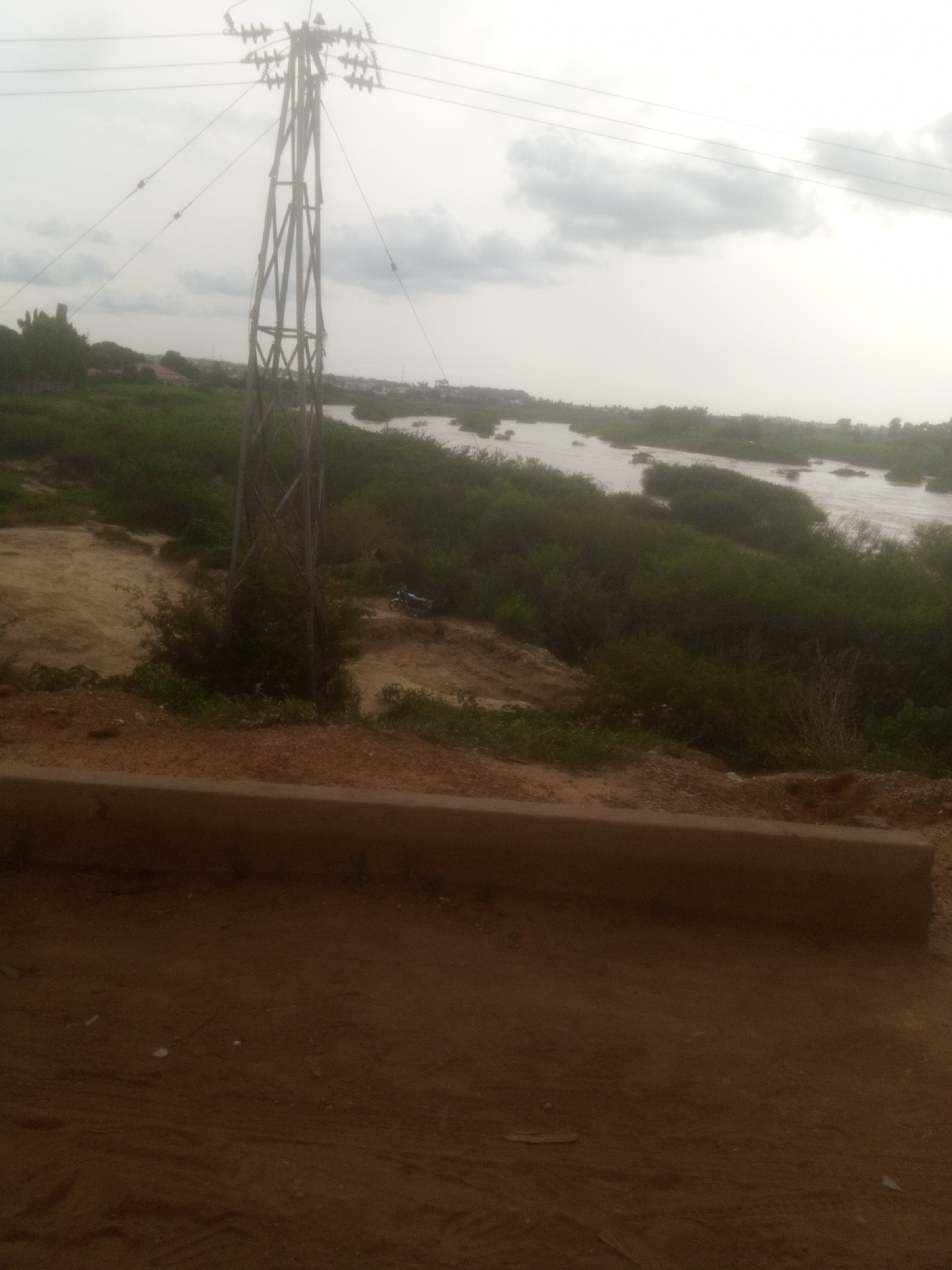 This is a full-frame, eye-level, outdoor, medium shot of a wide, shallow river with several small islands covered in lush green vegetation. The river appears to be in a flood state, with water flowing rapidly around the islands. In the foreground, there is a dirt path with a low, brown concrete barrier running horizontally across the frame. Behind the barrier, more dirt and scattered green vegetation are visible. To the left of the frame, a tall, metal electricity pylon stands prominently, with multiple wires extending outwards and upwards into the overcast sky. In the middle ground, behind the pylon and along the riverbank, dense green foliage lines the water. A dark-colored motorcycle is parked on the dirt path behind the pylon. The background shows a hazy, distant treeline and structures suggesting a settlement. The overall environment suggests a rural or semi-urban area near a body of water in Nassarawa, Nigeria, under a cloudy sky.