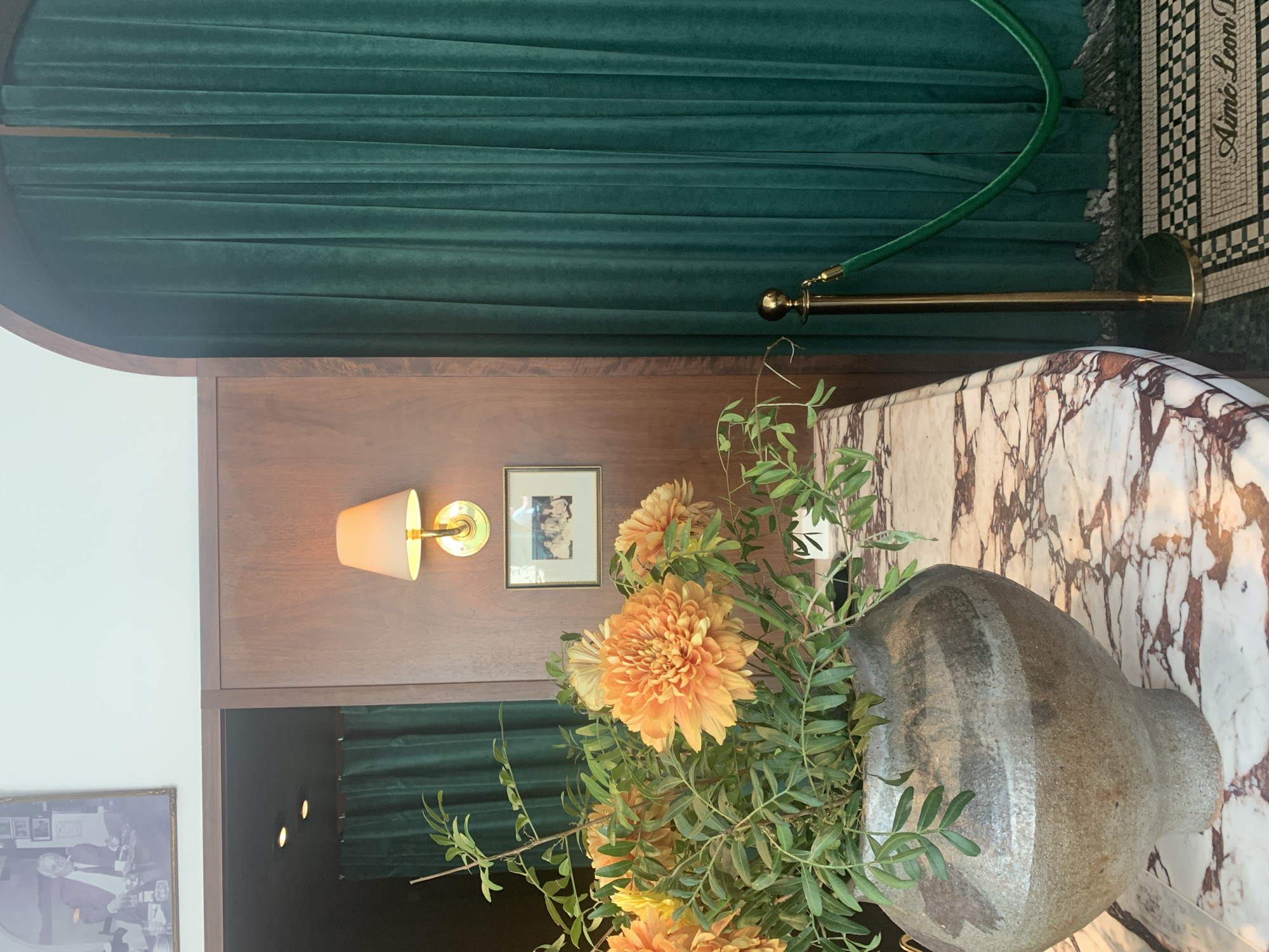 This media file captures an elegant interior setting, likely a high-end establishment such as a restaurant, cafe, or retail space in London, United Kingdom. The overall scene exudes a luxurious and sophisticated ambiance.
In the foreground, a large, rustic grey or charcoal-colored ceramic vase holds a bountiful arrangement of vibrant orange and yellow dahlia-like flowers, complemented by delicate olive branches. This floral display sits prominently on a polished marble counter, which features distinctive white, reddish-brown, and grey veining.
Behind the counter, a section of the wall is clad in warm, dark wooden paneling. A classic brass wall sconce with a cream-colored shade provides a soft, warm glow, illuminating two small framed pictures hanging on the wood. Below the wooden paneling, a darker recess reveals subtle recessed lighting and a hint of more deep green velvet curtains. A corner of another framed picture is visible in the bottom-left.
The background is largely defined by plush, dark green velvet curtains, draped richly to create a dramatic and opulent backdrop. To the right, a brass stanchion with a matching green velvet rope is positioned, suggesting a controlled entrance, queueing area, or demarcation of space. The flooring is a striking black and white mosaic tile, upon which the text "Aimé Leon D" is clearly visible, serving as a distinct location cue for an Aimé Leon Dore establishment.
No people are present in the image. The warm, inviting lighting from the sconce and the overall decor suggest an atmosphere suitable for an evening setting, or an interior designed to feel cozy and refined regardless of the time of day.