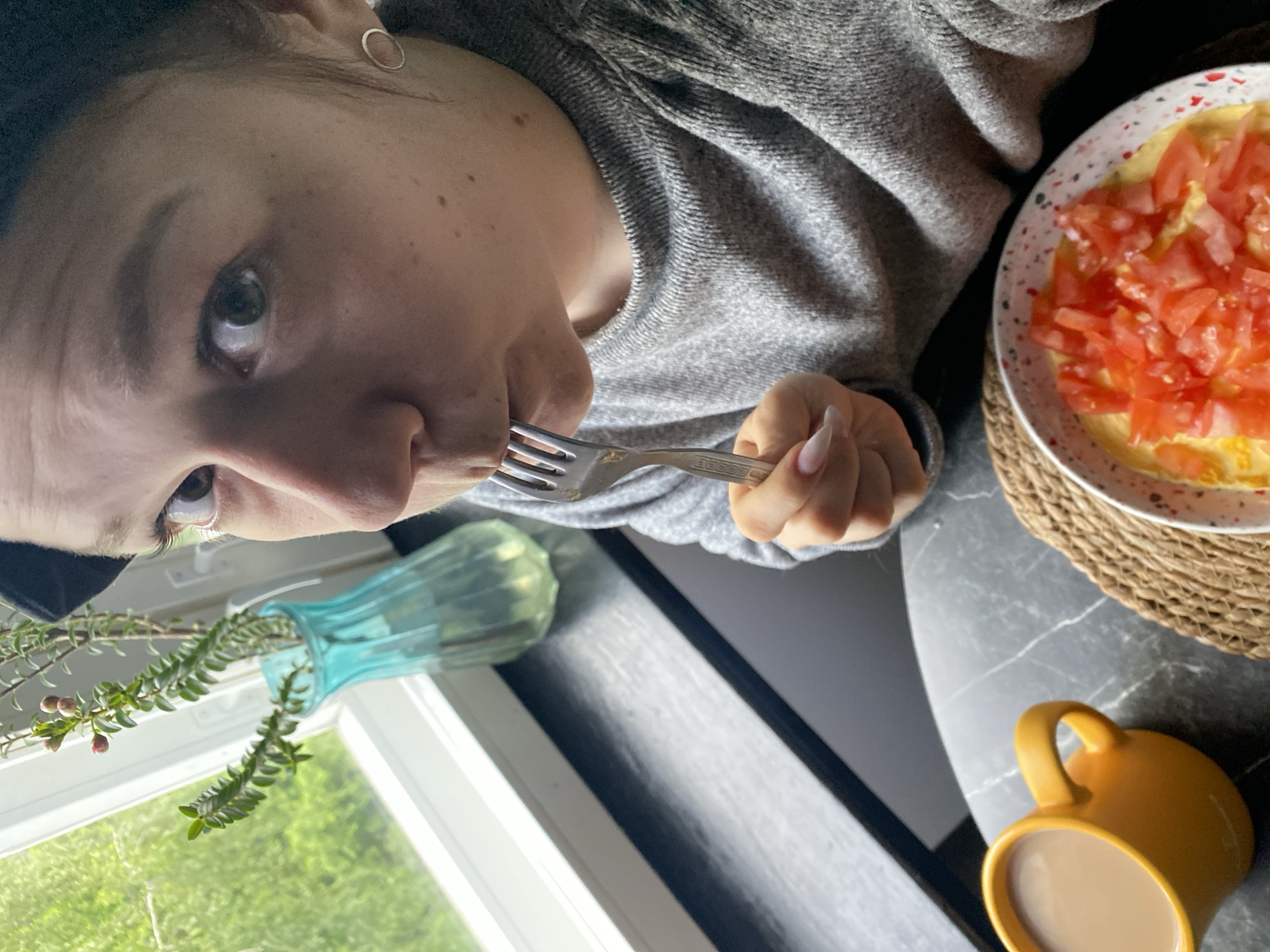 A woman is photographed in an upside-down orientation in an interior setting. She is holding a fork to her mouth, presumably eating. A plate of food, topped with diced tomatoes, rests on a woven placemat nearby. A yellow mug filled with a beige liquid is also visible. A light blue vase with greenery stands near a window, offering a view of outdoor greenery. The location is not discernible from the image.