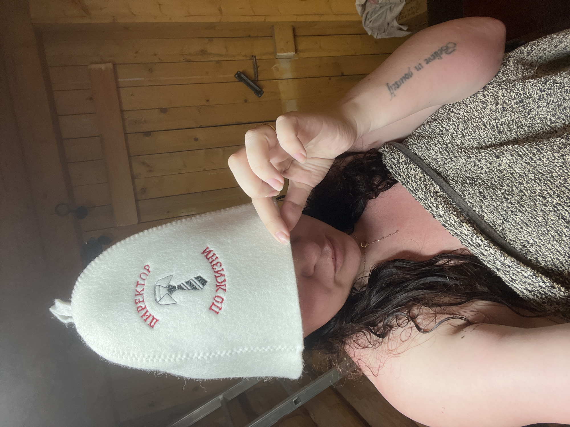A person lies on a wooden bench inside a sauna-like room with wooden plank walls. The individual is wearing a white sauna hat with Russian text and an image of a hammer and tie in red and black, partially obscuring the face. The individual is covered with a grey and white patterned towel or robe. An arm with a tattoo is visible above the robe. A small metal object is visible on the wall. The location is assumed to be a sauna in Valevaya, Russia.