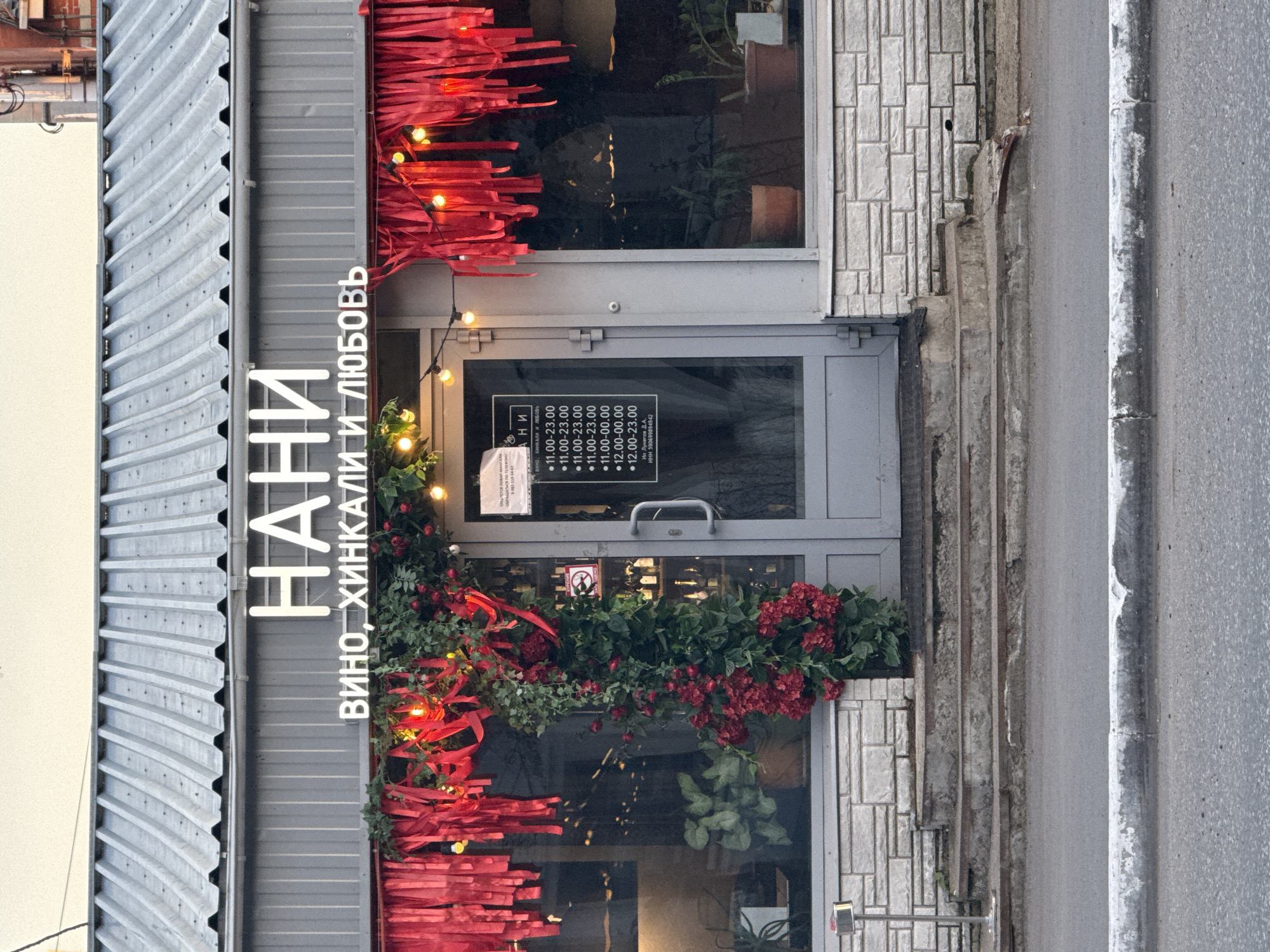 The image shows the facade of a business named "НАНИ" (NANI) in Votkinsk, Russia. The storefront features a prominent sign with the name in large, illuminated letters, and below it, "ВИНО, ХИНКАЛИ И ЛЮБОВЬ" (WINE, KHINKALI, AND LOVE). The entrance is a double door, with one half a door and the other a window. The facade is decorated with red streamers and string lights, and a large floral arrangement of green foliage and red flowers arches over the entrance. A sign on the glass door displays operating hours, indicating it is open from 11:00 to 23:00 or 00:00 on different days. The building has a corrugated metal roof and a facade clad in white brickwork. Stone steps lead up to the entrance from a paved sidewalk. The overall scene is one of a retail establishment, likely a restaurant or wine bar, with festive decorations.