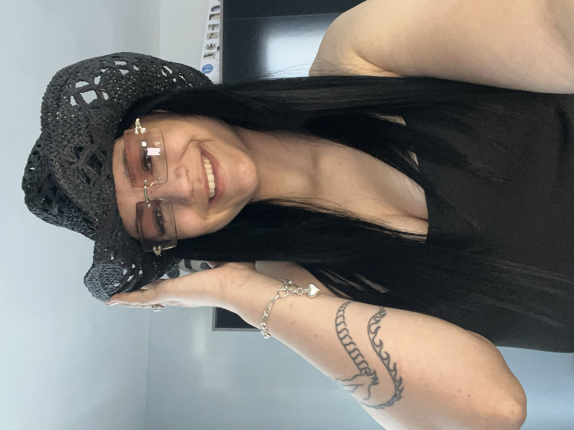 A woman with long, dark hair wears a black, crocheted hat and rimless sunglasses. She is smiling and looking towards the camera. A silver bracelet with a heart-shaped charm adorns her wrist, and she has a tattoo on her arm. The scene appears to be indoors, with a plain wall in the background. The lighting suggests it is daytime.