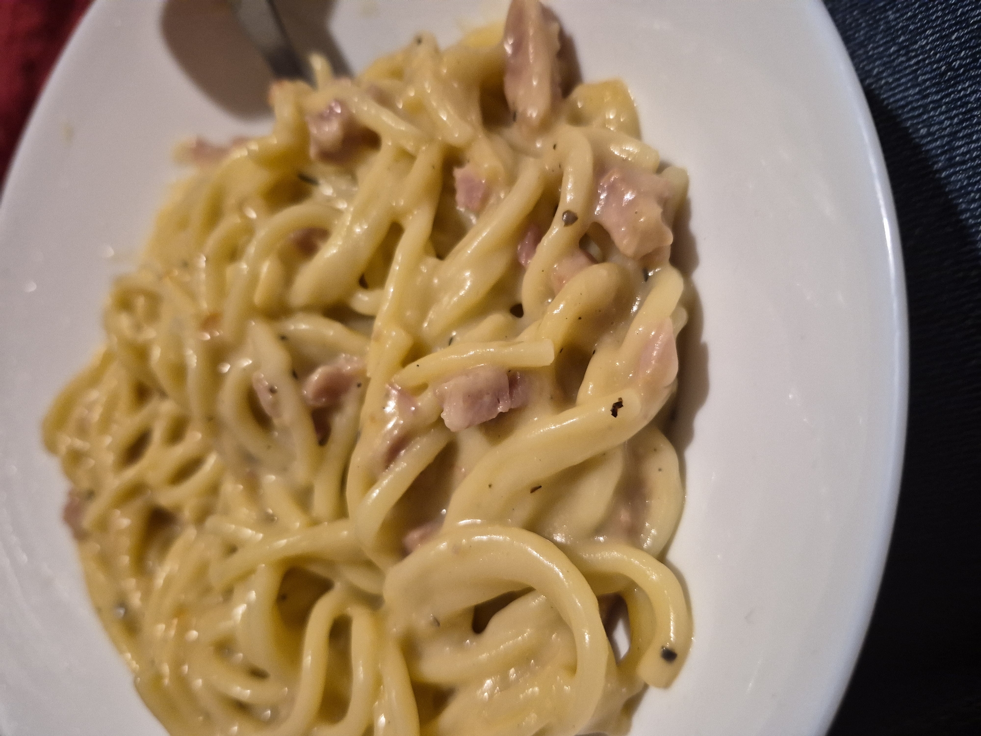 The image shows a close-up view of a plate of spaghetti carbonara. The pasta is coated in a creamy sauce and mixed with pieces of pancetta or bacon. Black pepper is visible on the pasta. A fork can be seen on the right side of the plate, suggesting the dish is being eaten. The plate is white and rests on a dark surface, possibly a table. The background is out of focus, but appears to be a dark reddish color. There are no people visible in the image, and the setting is a domestic environment. There is no indication of location in Sandwell, United Kingdom.