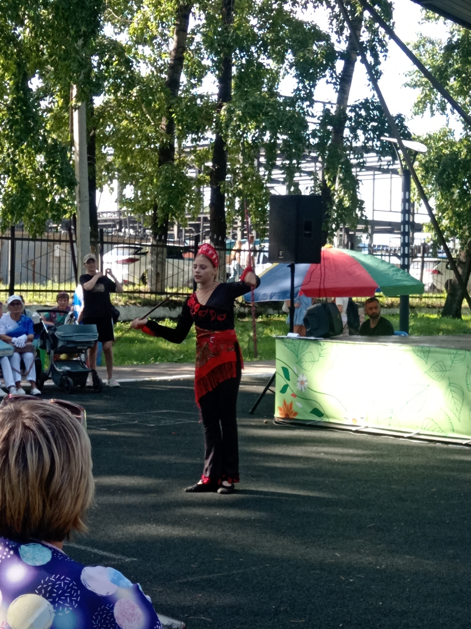 A young girl in a black and red costume, with a red headpiece, is performing outdoors, holding a long, thin object. She is standing on a dark, speckled surface, possibly asphalt or rubberized flooring. Behind her, a man in a black t-shirt and a baseball cap is visible, possibly filming or taking pictures. Further back, there are several people seated, including two women in light-colored clothing and a child in a stroller. A portable speaker is set up to the left of the performer. A table draped with a green cloth decorated with foliage and flowers is set up behind the speaker and to the right. Partially visible behind the table are two multi-colored umbrellas providing shade, and a man with a beard sitting behind the table. The background shows trees with green leaves and a metal fence, suggesting an outdoor public space. The overall scene appears to be a performance or event taking place during the day. The scene is consistent with an outdoor public event in the city of Belogorsk, Russia, given the context provided.