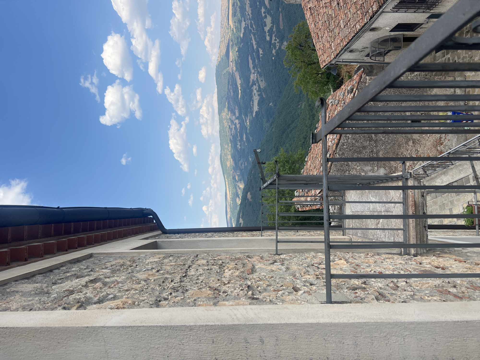 The image captures a view from a balcony in Viggiano, Italy, looking out over a valley. The foreground features a stone wall and a metal railing of a balcony. To the right, another building with a terracotta tiled roof is visible. Beyond the buildings, rolling green hills dominate the landscape, stretching towards a distant mountain range under a bright blue sky dotted with white clouds. The scene appears to be a quiet, scenic rural or semi-rural setting within the town of Viggiano, likely showcasing its characteristic hilly terrain and traditional architecture. There are no people or animals visible in the frame.