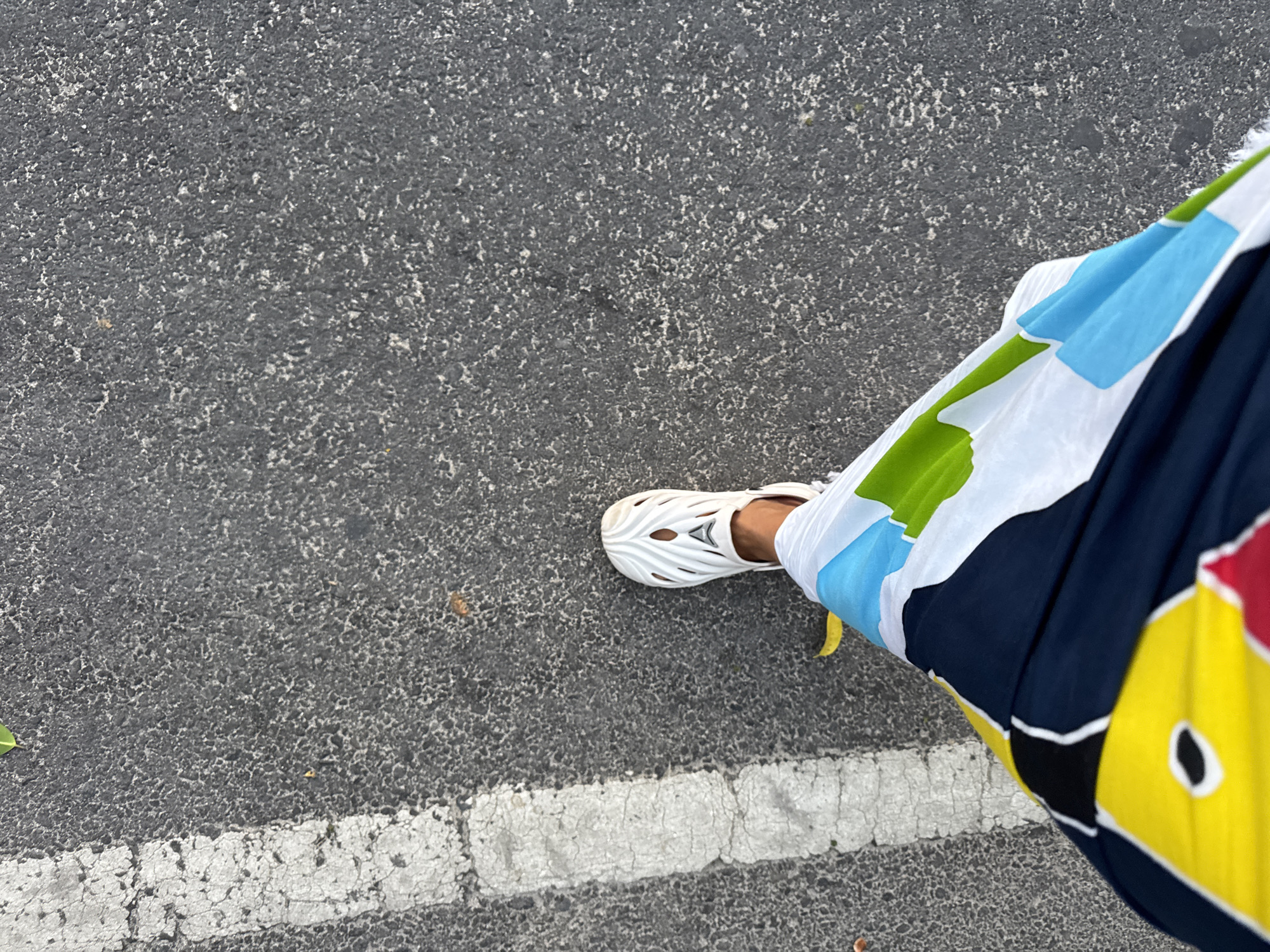 A downward-facing shot taken on an asphalt surface, possibly a street, depicts a person's leg and foot adorned with a white, sandal-like shoe with cut-out designs. The person is wearing a brightly colored garment featuring shades of blue, green, white, yellow, red, and black. A white line is visible on the asphalt, situated near the lower left corner of the frame. The location appears to be Jimbaran, Indonesia.