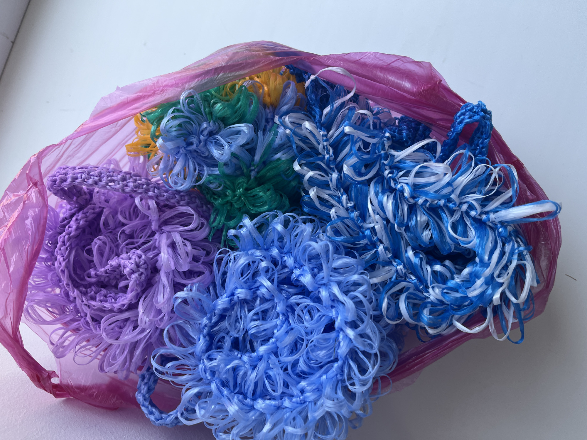 A pink plastic bag contains several multi-colored crocheted items. The items are made from what appears to be plastic or synthetic string, and they are shaped like loose, floral or pom-pom like forms. Colors include purple, blue, green, yellow, and white. The bag and its contents are placed on a white surface, with a pale, textured wall in the background. This scene is not in Blagoveshchensk, Russia.