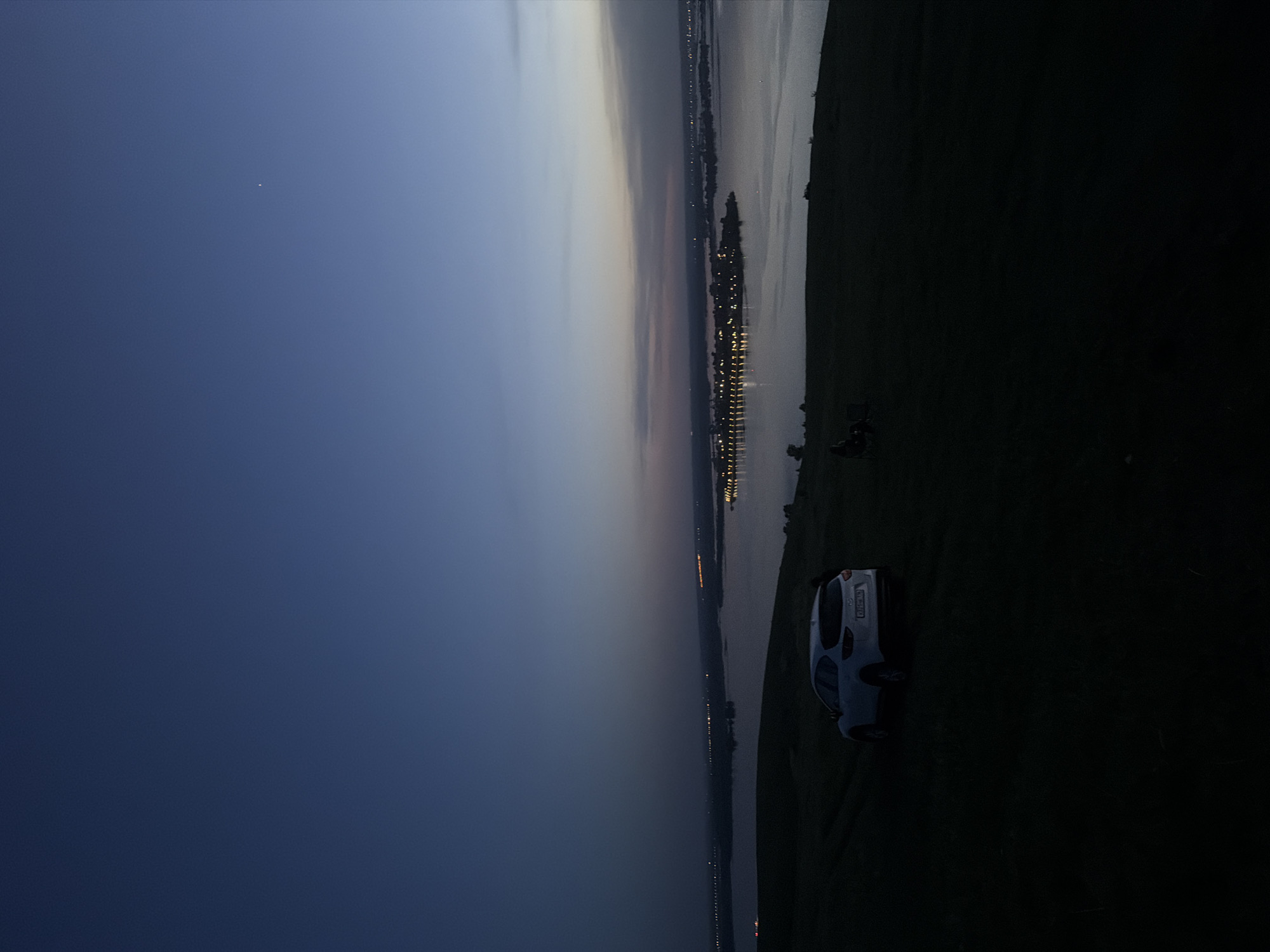 A nighttime, landscape-oriented image displays a view from a high point, likely a hill or slope, overlooking a body of water. A small white car is parked on the grassy terrain in the foreground, slightly right of center. A bicycle is also visible near the car. Across the water, a city skyline is illuminated with scattered lights stretching along the horizon. The sky transitions from dark blue to lighter shades of gray and pink, indicating either dusk or dawn. A single bright point of light, possibly a star or a distant light source, is visible in the dark blue sky. The location is inferred to be near the city of Innopolis, Russia, based on the prompt. The overall scene evokes a sense of tranquility and vastness.