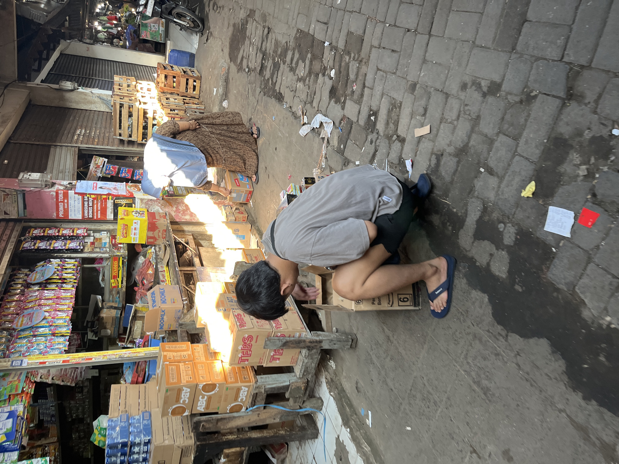 The image captures a street scene in Larangan, Indonesia, likely a market area. In the foreground, a young man wearing a grey t-shirt and black shorts is crouched on the ground, facing away from the camera. He is wearing blue flip-flops. Next to him, a stack of cardboard boxes is visible, one of which has the number "6" printed on it. To the left, more cardboard boxes are stacked, some labeled with "ABC" and "TEBS". In the mid-ground, a woman wearing a brown patterned dress and a light blue headscarf is bending down, interacting with a cardboard box. Behind her, a section of a market stall is visible with various goods displayed. Wooden crates are stacked to her right. The ground is paved with cobblestones and appears wet in places, with debris scattered around. The background shows more market stalls and structures, with a motorcycle parked on the right side of the image. The lighting suggests it is daytime, with sunlight casting shadows.