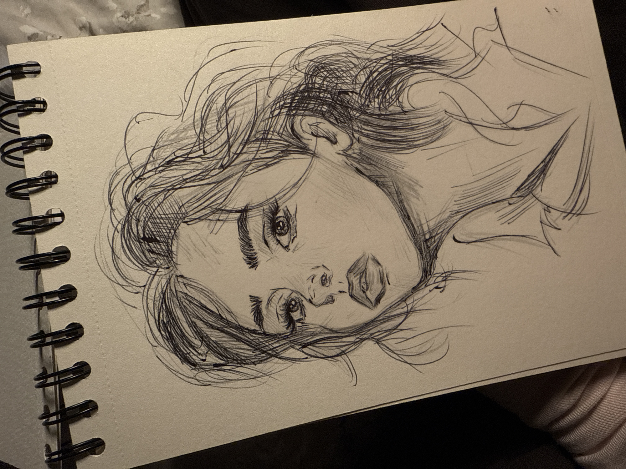 The image is a sketch of a person's head and shoulders on a page in a spiral-bound sketchbook. The person in the sketch has long, loose hair. The sketch is in pen or pencil, and the style is loose and expressive.
The image contains no information that could be used to pinpoint a location in Sofia, Bulgaria, or any city for that matter. There are no contextual landmarks, signs, or other location-specific details.