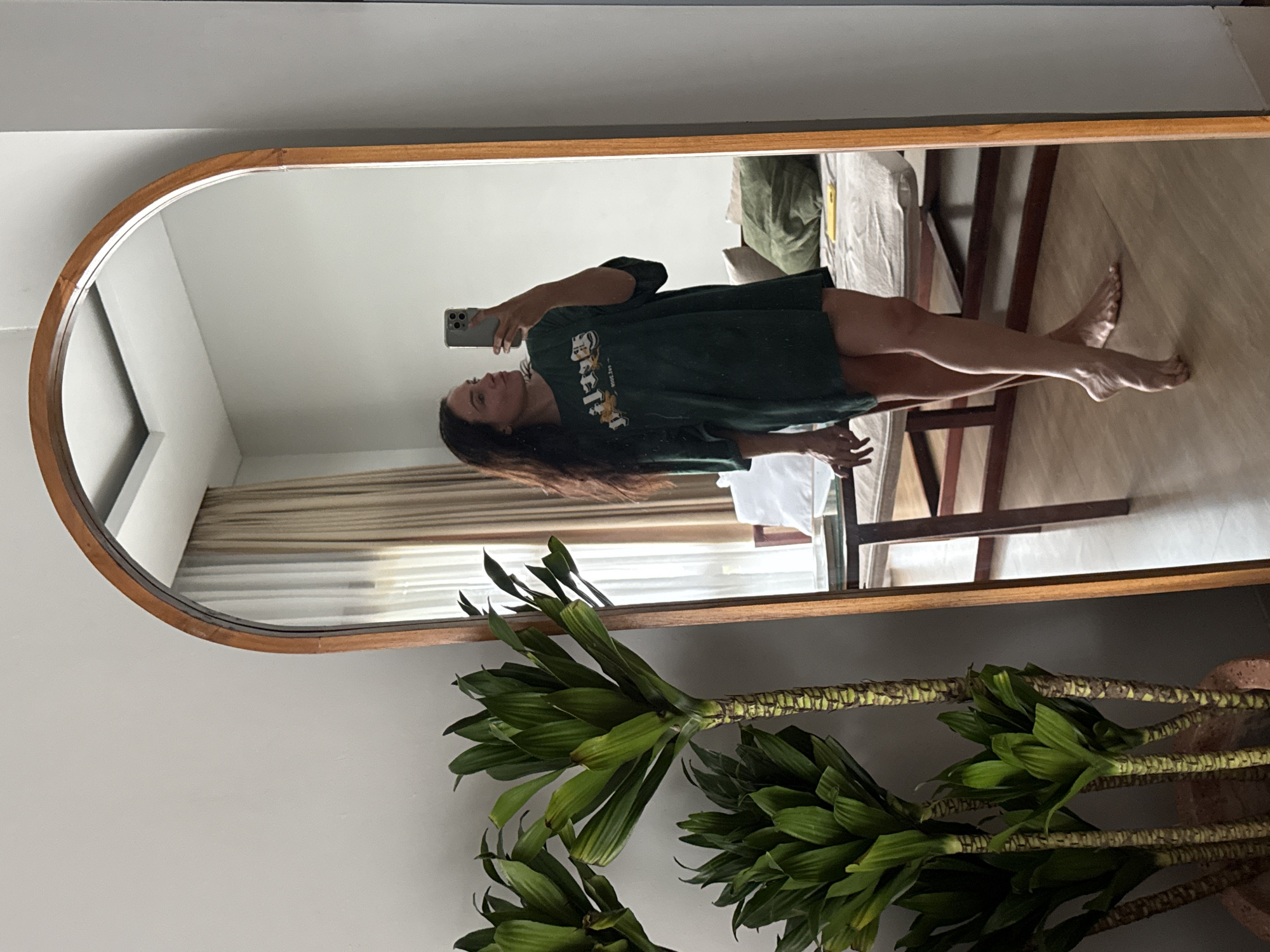 A full-body mirror reflects a woman standing and taking a selfie in a room in North Kuta, Indonesia. The woman, with long dark hair, is wearing a loose, dark green t-shirt that extends to her mid-thighs. She is holding a smartphone in her right hand. The room appears to be a bedroom, with a bed visible in the background. The bed has a light-colored comforter and several pillows, one of which is a large green cushion. A wooden bedside table with a dark wooden frame is also present. To the left of the mirror, a large potted plant with green, sword-shaped leaves is visible. The mirror itself has a rounded top and a wooden frame. The lighting in the room is soft, suggesting it may be daytime.