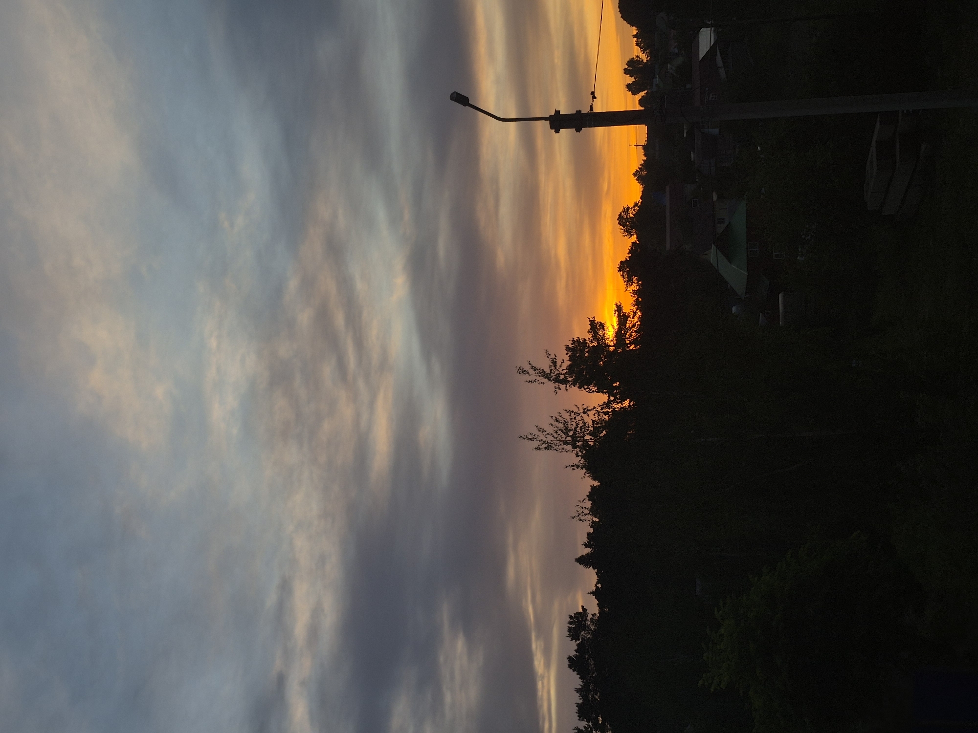 The image captures a twilight scene in what appears to be Chaikovsky, Russia. The sky displays a gradient from gray and blue on the left to a vibrant orange near the horizon, indicating a sunset or sunrise. A silhouetted treeline and houses are visible against the orange sky. A utility pole stands prominently on the right side of the image. The foreground is dark and obscured, primarily consisting of foliage and indistinct objects. No people are directly observable in the scene.
