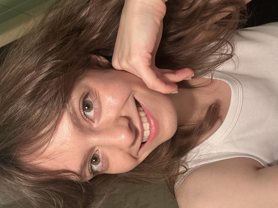 A close-up indoor portrait captures a young adult female. She possesses light skin, brown hair with bangs, and light green eyes, looking directly forward with a wide smile revealing upper and lower teeth. Her right hand is positioned against the right side of her face, with fingers slightly curled. She is dressed in a white ribbed tank top. The background is uniformly dark and featureless. No specific geographic identifiers, including a location in Moscow, Russia, are discernible from the image.