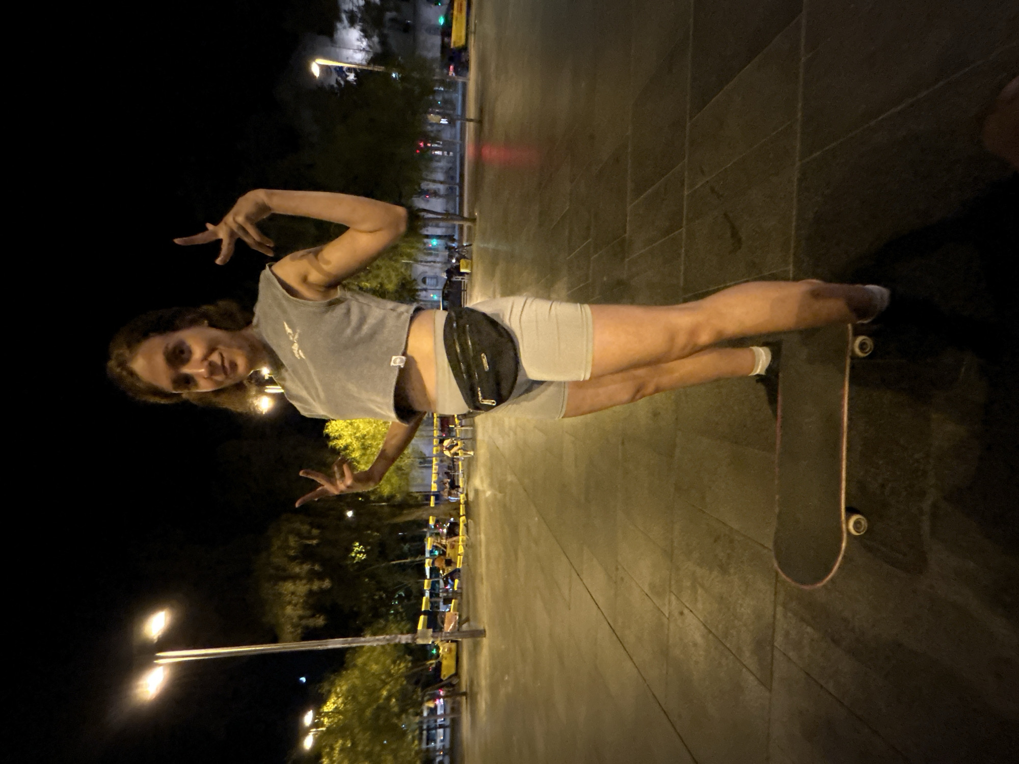A person is standing on a skateboard in a public square at night, likely in Barcelona, Spain, as suggested by the presence of yellow taxis and the general urban night scene. The person is wearing a grey cropped t-shirt and grey shorts, with a black fanny pack around their waist. They are making a peace sign with their right hand and a different hand gesture with their left hand, looking towards the camera with a slight smile. The ground is paved with large square tiles. In the background, there are streetlights, trees, and some buses and cars. The overall lighting suggests it's a well-lit urban area at night.