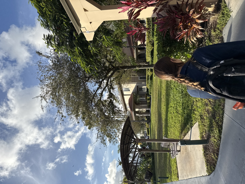 A woman with a blonde braid wears a navy shirt and black backpack. A house is visible in the background with a brown roof and white pillars. Trees with bare branches spread out in the yard. Lush green grass covers the ground. Part of a metal structure with benches sits to the left of the scene, facing toward the house. The sky is blue with scattered white clouds. Red tropical plants are in the corner.
