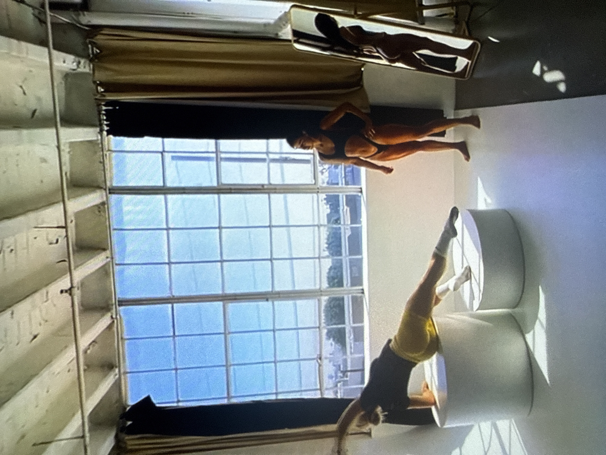 The scene depicts an indoor studio space situated in the city of Varna, Bulgaria, illuminated by natural light entering through large, multi-pane windows that reveal a distant cityscape and trees under a clear blue sky. The ceiling features exposed wooden beams and utility pipes. The floor is bifurcated, presenting a white surface on the left and a dark, possibly black, surface on the right.
Two individuals are present within the space. On the left, a person with dark hair, wearing a black short-sleeved top, yellow shorts, and white socks, is positioned dynamically across two white cylindrical platforms of varying heights. This individual supports their weight with their left arm on the taller platform while their body extends diagonally, with their right leg resting on the shorter platform. On the right, a second individual with dark hair, barefoot and wearing black two-piece swimwear (a top and a high-cut bottom), stands facing slightly left, looking towards the window. A tall, rectangular mirror with a gold frame stands upright on the dark floor to the right, reflecting the profile of the second individual. Dark and brown curtains are visible framing the large window on either side. Strong natural light from the window casts pronounced shadows from the subjects and objects onto the floor.
