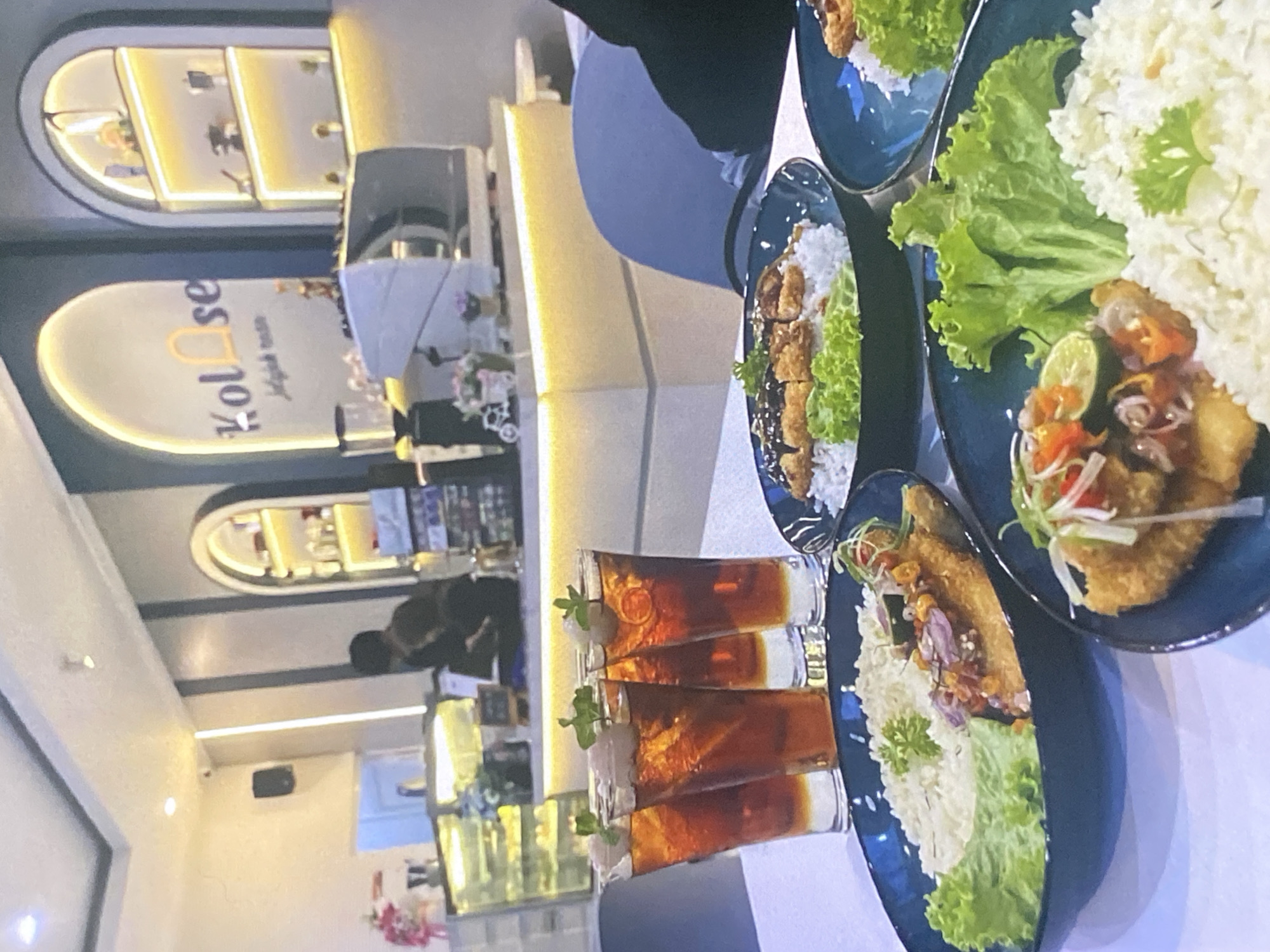 In the city of Tebel, Indonesia, a restaurant scene unfolds featuring several plates of food and beverages. Four tall glasses of iced tea, each garnished with a slice of lychee and a sprig of mint, stand prominently on a white table. Adjacent to the drinks are three dark blue bowls filled with rice, topped with fried chicken cutlets and a garnish of lettuce and chili sauce. The background reveals a modern cafe interior with arched alcoves displaying merchandise and illuminated shelves. A coffee machine and grinder are visible on a counter, suggesting the establishment's offerings. The ambiance is bright and contemporary, with a soft yellow glow emanating from the decorative lighting.