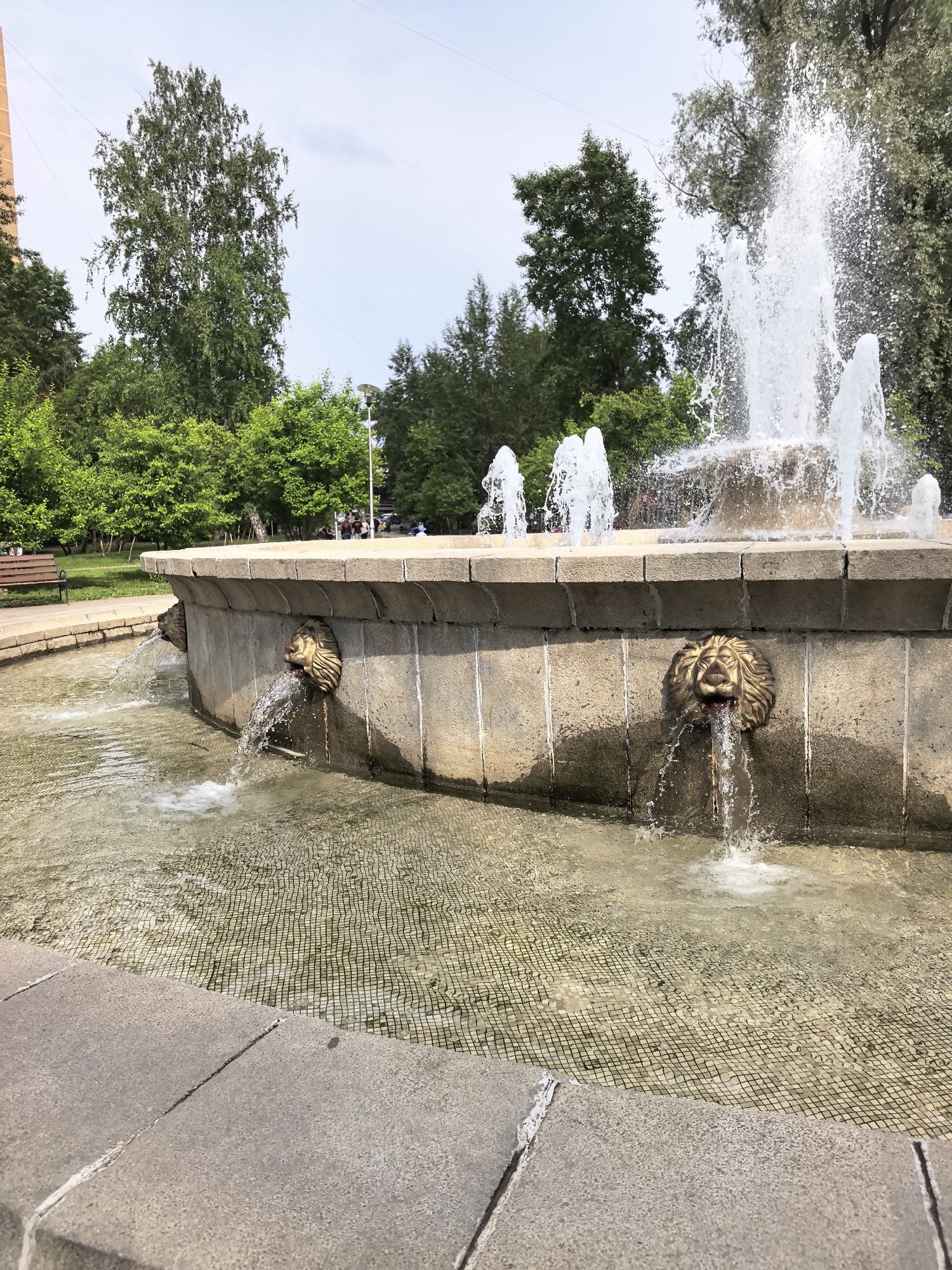 The image displays a lively fountain in a public park or square, captured on a bright day. The central subject is a large, circular fountain constructed from light-colored stone or concrete. Multiple powerful jets of water spray high into the air from the fountain's upper tier. Along the outer wall of its lower tier, several ornate, bronze-colored lion-head spouts discharge streams of water into the shallow basin below. The bottom of the fountain's pool is visible through the clear water, revealing a distinctive tiled or patterned surface.
Surrounding the fountain are numerous lush green trees, some with dense foliage, suggesting a well-maintained park environment. To the left, a wooden park bench is visible, nestled among the greenery. In the background, beyond the trees, a few indistinct figures of people can be seen, indicating the presence of other visitors. A street lamp is also visible among the trees on the right. The scene is brightly illuminated by natural daylight under a partly cloudy sky, suggesting a pleasant morning or afternoon. The overall atmosphere is serene and inviting. There is no visible text in the image.