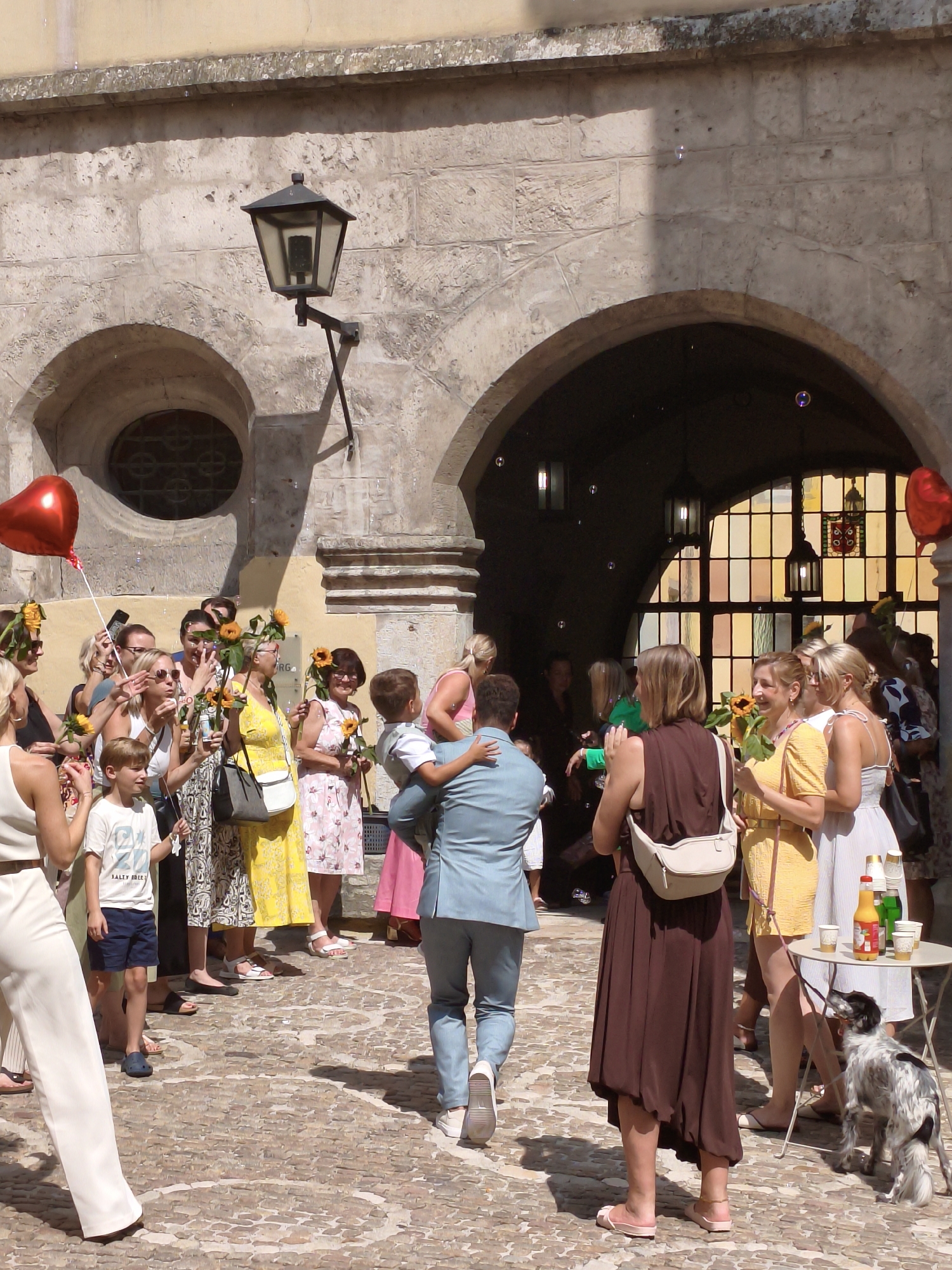 The image shows a group of people gathered outdoors in what appears to be a courtyard or public square. The architecture suggests a historic European city, with stone buildings, arched doorways, and cobblestone paving. The scene is bathed in bright sunlight, and there are several bubbles floating in the air, indicating a festive occasion, possibly a wedding.
In the foreground, a man in a light blue suit is carrying a young boy who is looking away from the camera. To their left, a woman in a brown dress with a white crossbody bag is looking towards the man and boy. Behind them, a group of people, mostly women, are holding sunflowers and some are taking photos. One woman is holding a red heart-shaped balloon on a string.
The background features a large arched opening leading to a dimly lit interior with several pendant lamps and a stained-glass window. To the right, a small round table is set up with cups and bottles, and a black and white dog is standing nearby, looking at the table.
While the specific location in Regensburg, Germany is not explicitly identified in the image, the architectural style and the presence of cobblestone streets are consistent with the historic old town of Regensburg. The building appears to have a cloister or a similar medieval structure. The overall atmosphere is celebratory and joyful.
