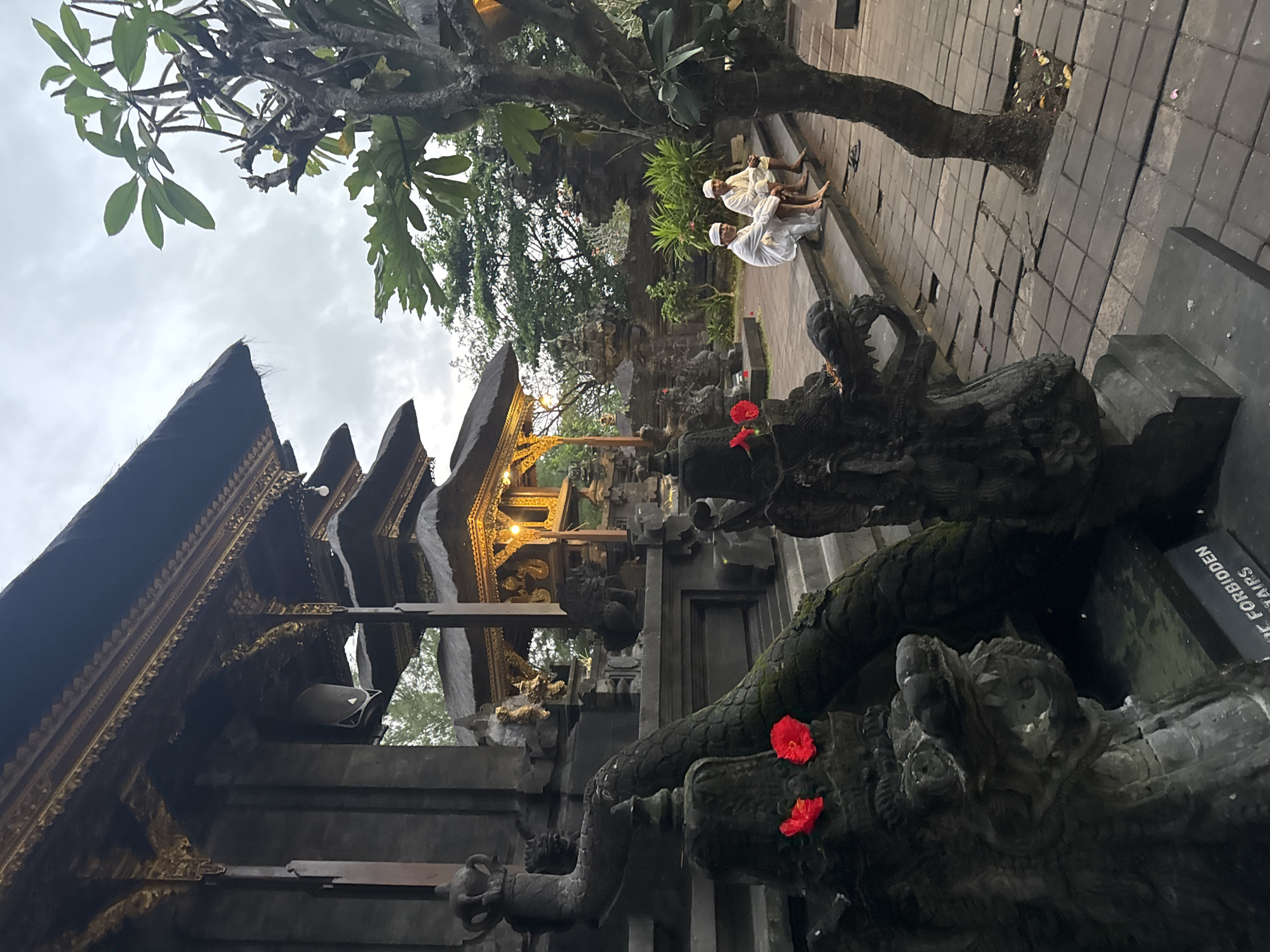 The image depicts a scene at a temple in Dawan, Indonesia. In the foreground, two dark stone dragon statues with red flowers on their heads are visible, one with its mouth open. To the right, two men in white traditional clothing are sitting on stone steps. Behind them, the temple architecture features multiple tiers of dark, thatched roofs with ornate golden detailing. The ground is paved with square stones, some of which are cracked or uneven. Trees and lush greenery surround the temple, suggesting an outdoor setting within a temple complex. The sky is overcast, and the lighting indicates it may be dusk or a cloudy day.
