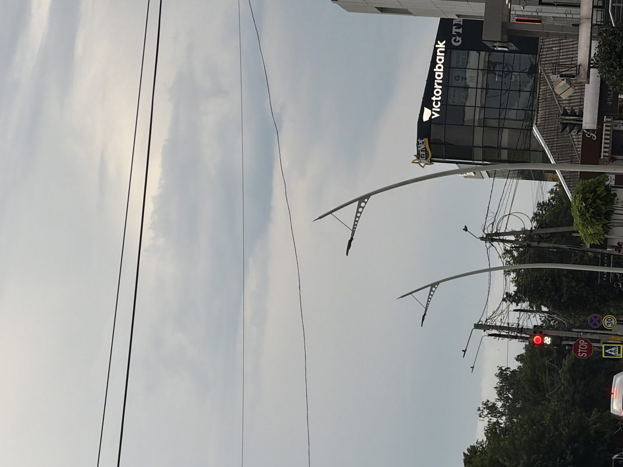 The image captures an urban scene on an overcast day in Strășeni, Moldova. A modern, glass-fronted building with the prominent "Victoriabank" logo is visible. Adjacent to it is a structure with "GTM" signage. Multiple utility poles with an array of wires crisscross the sky. Two contemporary streetlights with multiple illuminating points stand tall, their curved arms reaching upwards. In the lower left quadrant, a traffic light shows a red signal, accompanied by a "STOP" sign and a pedestrian crossing sign. Further down, a blue circular sign with the number "30" indicates a speed limit. Trees line the background, and a car is partially visible in the lower left corner. The overall impression is of a typical street view within a developed area.