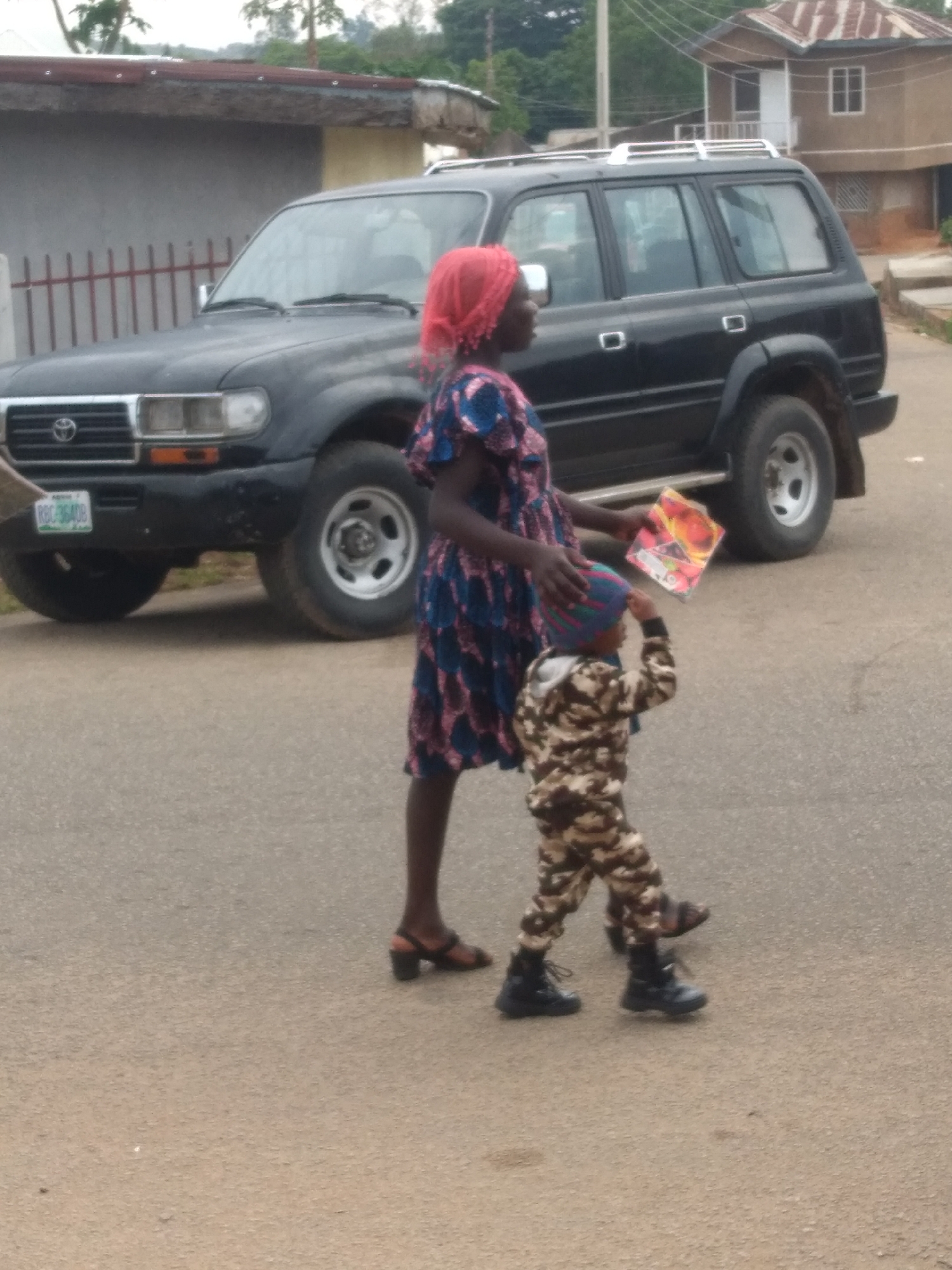 An adult and a child are walking on a paved, light grey road in an outdoor, semi-residential setting. The adult, appearing to be a woman, is on the left, wearing a red head covering, a blue and pink patterned dress, and brown sandals with heels. She holds a colorful rectangular item, possibly a book or toy packaging, in her left hand, and her right hand is placed on the child's head. The child, a toddler or young preschooler, walks slightly ahead and to the right of the adult, wearing a multicolored knit hat, a camouflage-patterned long-sleeved top and pants set, and black boots. The child has their right arm raised towards their hat.
In the background, a dark grey Toyota Land Cruiser SUV (J80 series) with a silver roof rack is parked facing left. Its front license plate displays "RBC-364DB". Behind the SUV, a two-story building with a brown exterior and a rust-colored corrugated metal roof is visible, featuring white-framed windows and a white door. To the left of the SUV, a grey building with a reddish-brown roof and a low metal fence is partially visible. Green vegetation and trees are present in the far background. The sky appears overcast.