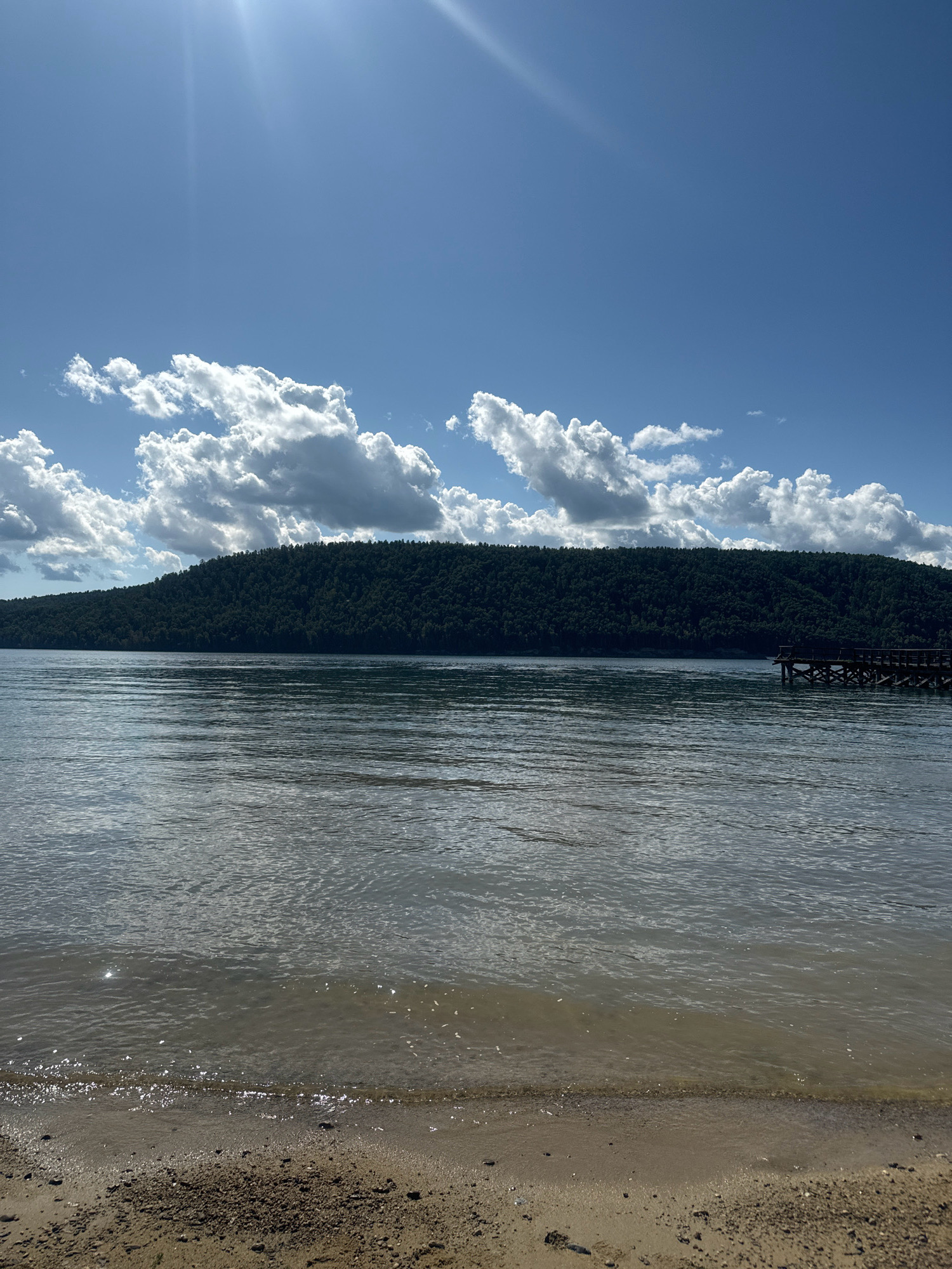The image depicts a serene natural landscape. In the foreground, a sandy shore meets a body of water that appears to be a lake or a large river. The water surface is calm with gentle ripples reflecting the sunlight. Across the water, a densely forested hill rises, its dark green foliage creating a rich texture. Above the hill, a bright blue sky is dotted with fluffy white clouds. The sun's rays pierce through the clouds, casting a bright sheen on the water and sky. To the right, a wooden pier juts out into the water, its dark structure contrasting with the surrounding natural elements. The scene suggests a peaceful, outdoor setting, possibly a recreational area or a natural preserve. The location is identified as Тальцы (Иркутский), Russia.