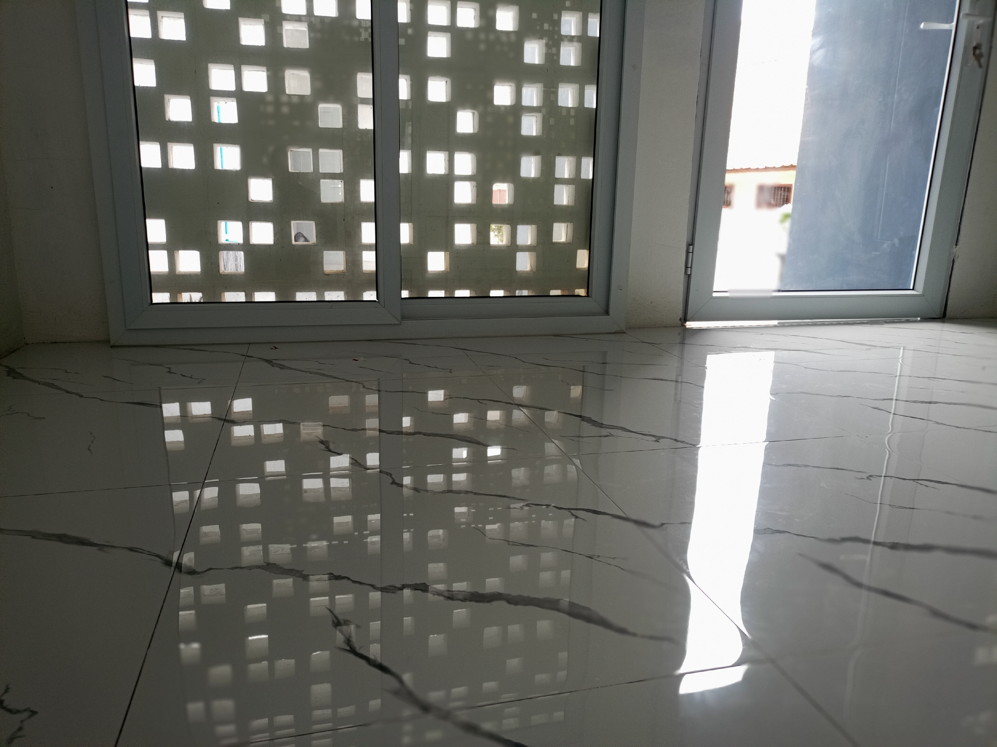 The image shows a glossy tiled floor reflecting sunlight and the pattern of a window with small square openings. A partially open door is visible in the background. The location is likely indoors in a building in Bireuen, Indonesia. No people are visible.
